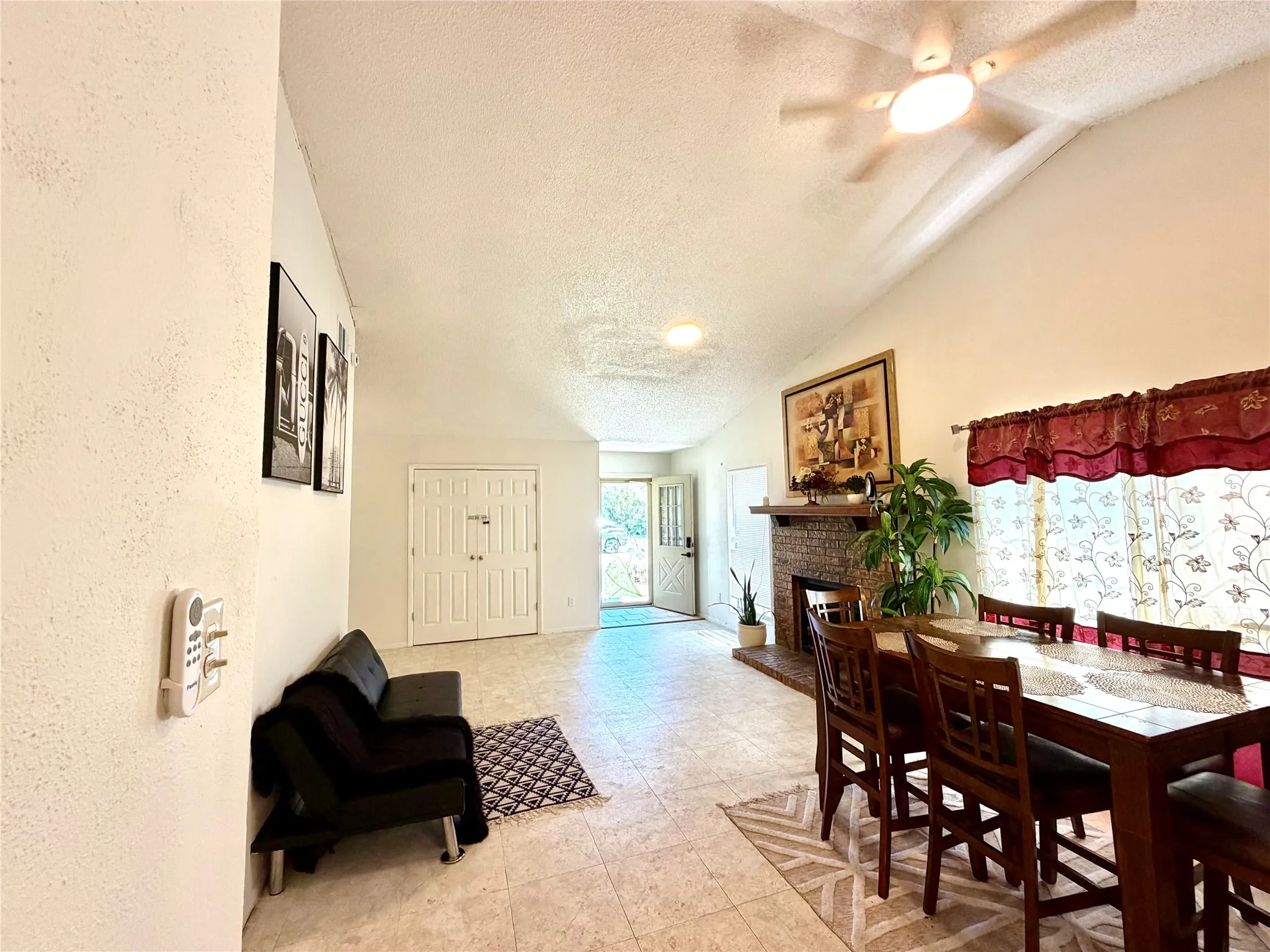 Living and dining area featuring vaulted ceilings, a ceiling fan with integrated lighting, a brick fireplace with a wood mantle, and light-toned tile flooring