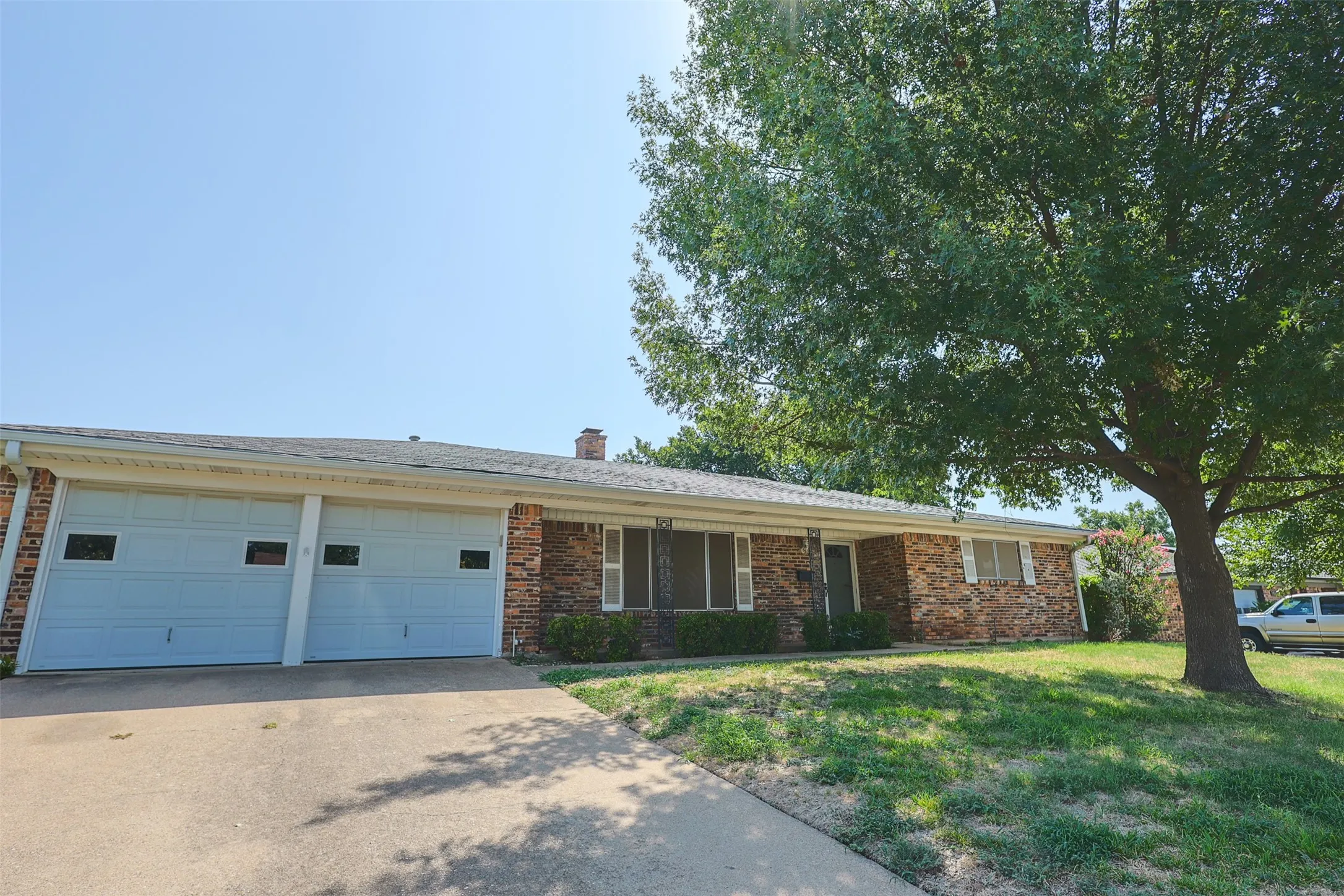 Brick facade residence featuring a two-car garage with panel doors, a concrete driveway, and a mature shade tree
