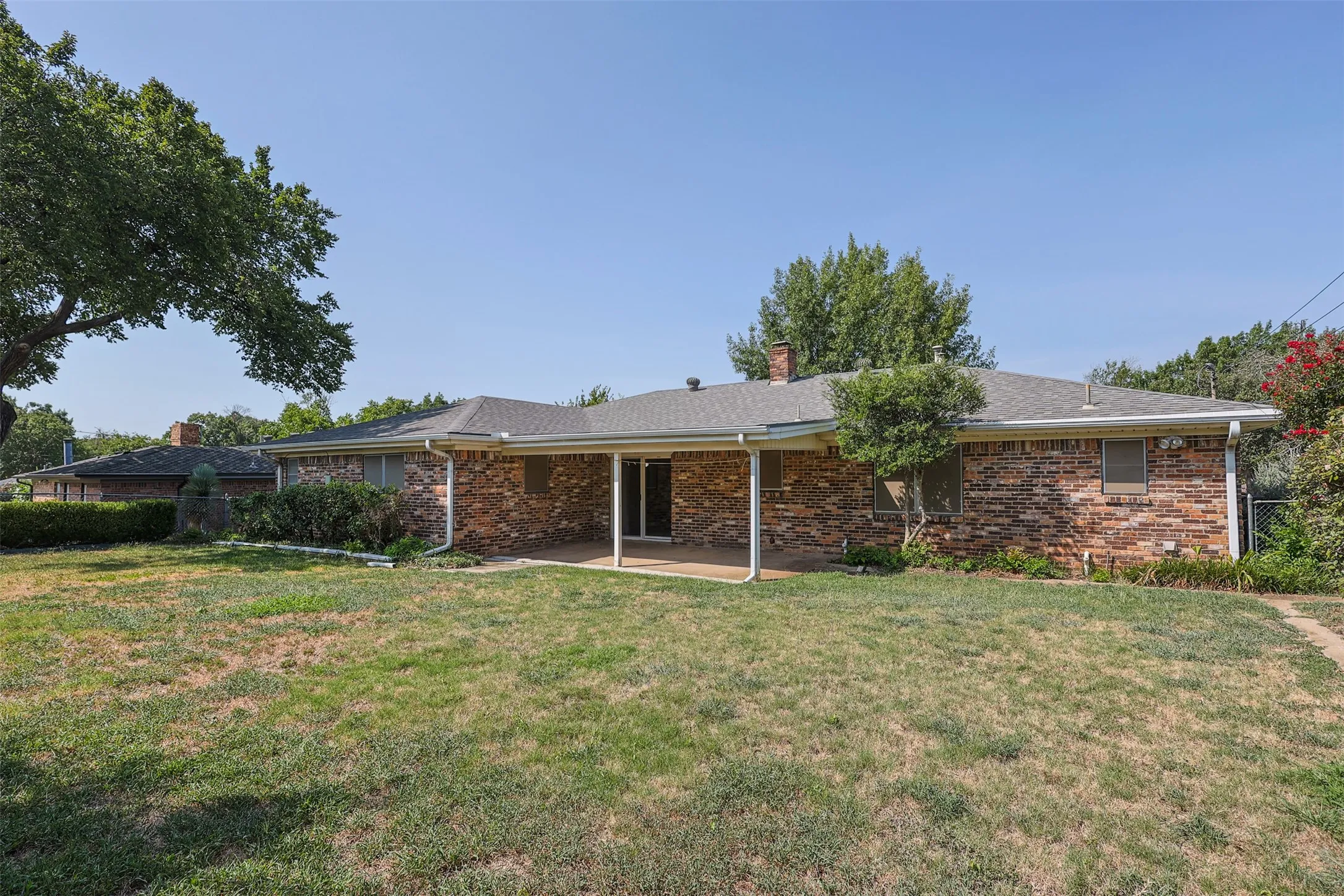 Expansive backyard featuring a covered patio, brick exterior, and a mature tree providing natural shade