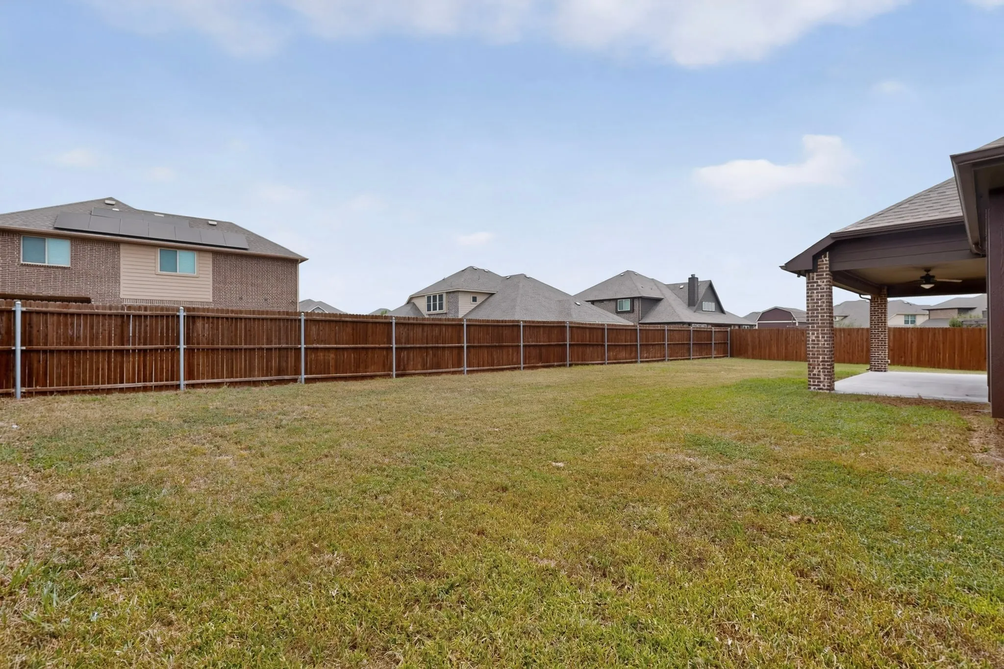 Expansive backyard featuring a full perimeter privacy fence, concrete patio, and covered outdoor living space with ceiling fan and brick-wrapped columns