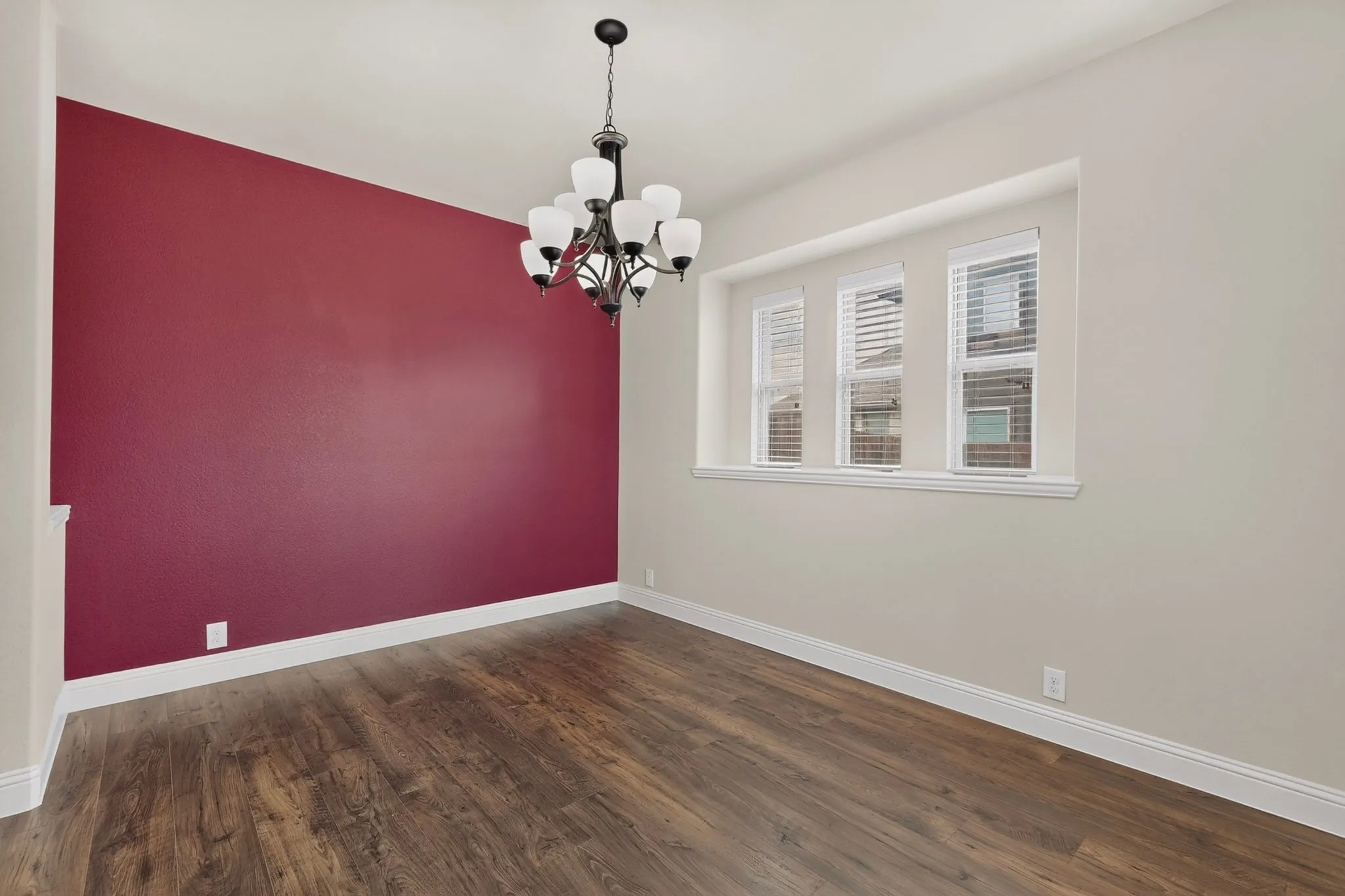 Spacious room featuring wood-finish flooring, a contemporary chandelier, a bold accent wall, and three windows with white blinds
