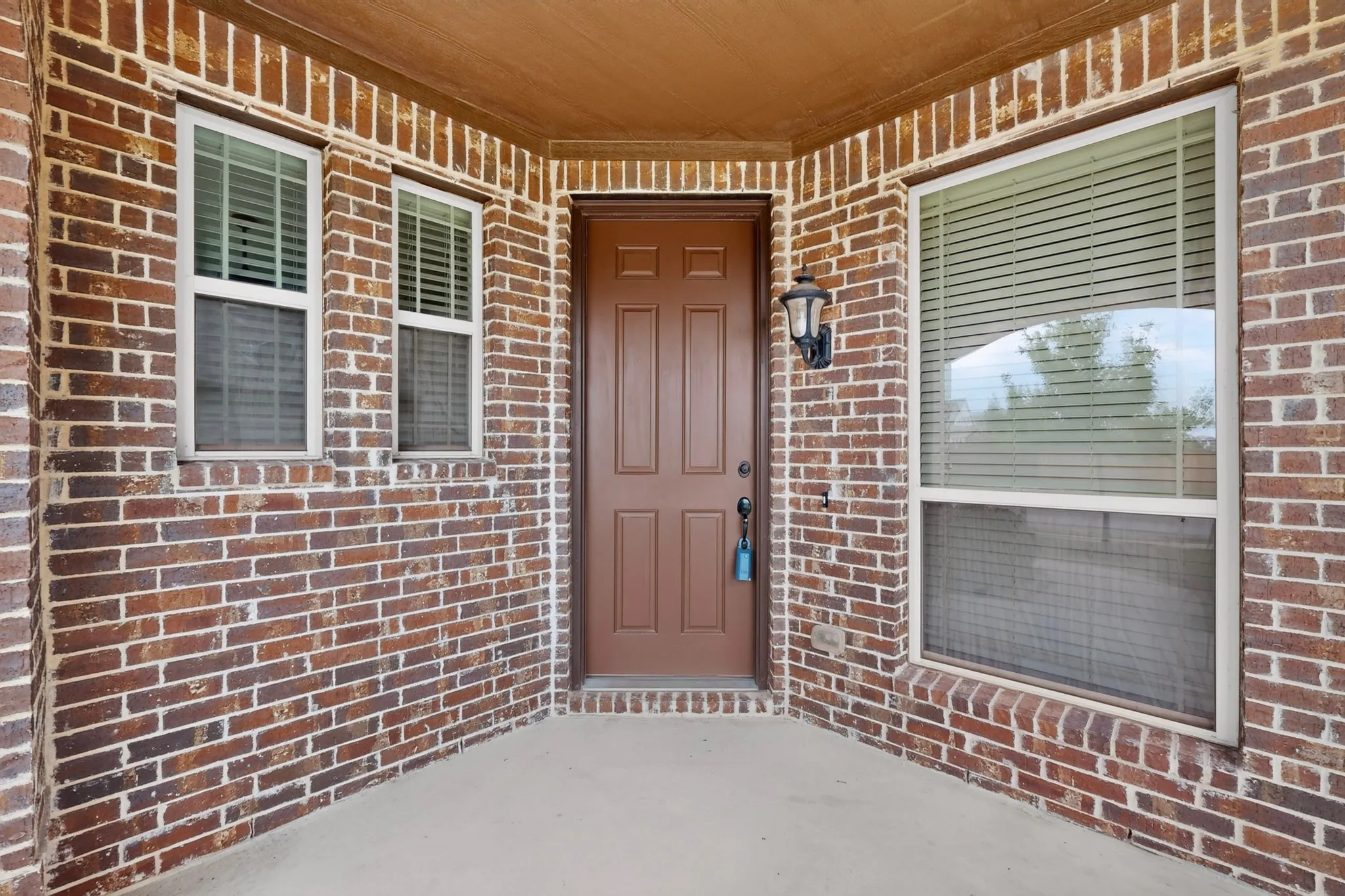 Brick exterior entryway featuring a paneled door, two fixed windows with blinds, an overhead soffit, and an outdoor wall sconce