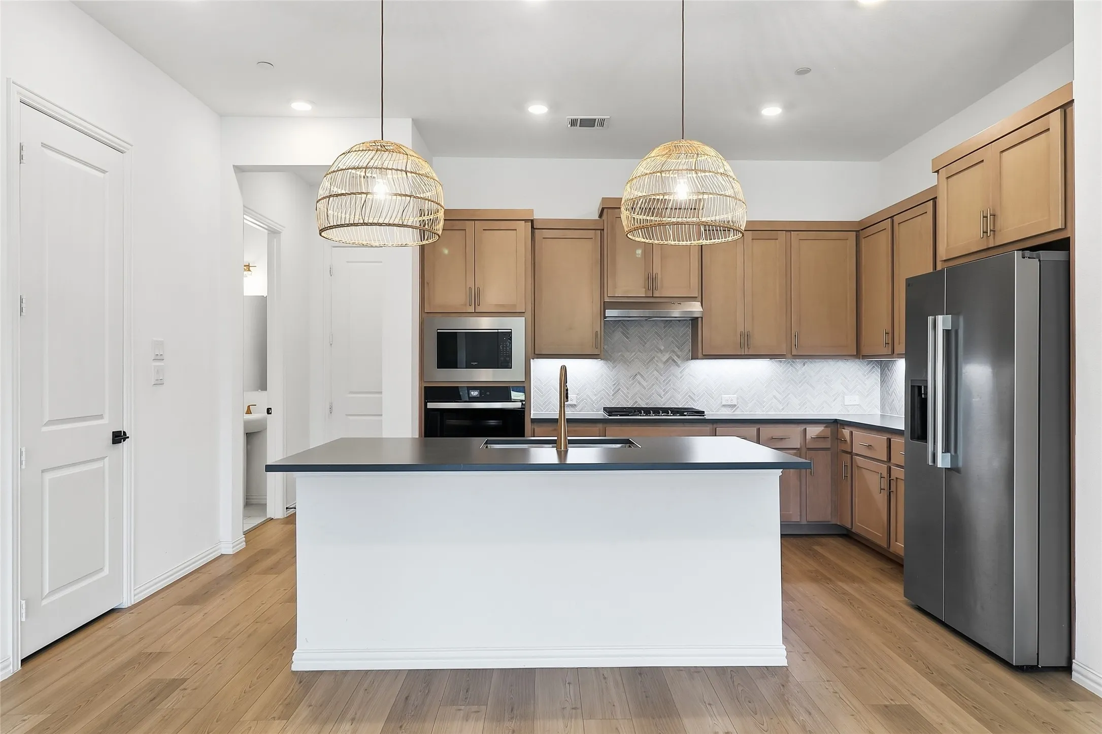 Kitchen featuring a central island with countertop, wood-finish flooring, stainless steel appliances, herringbone tile backsplash, and natural wood cabinetry