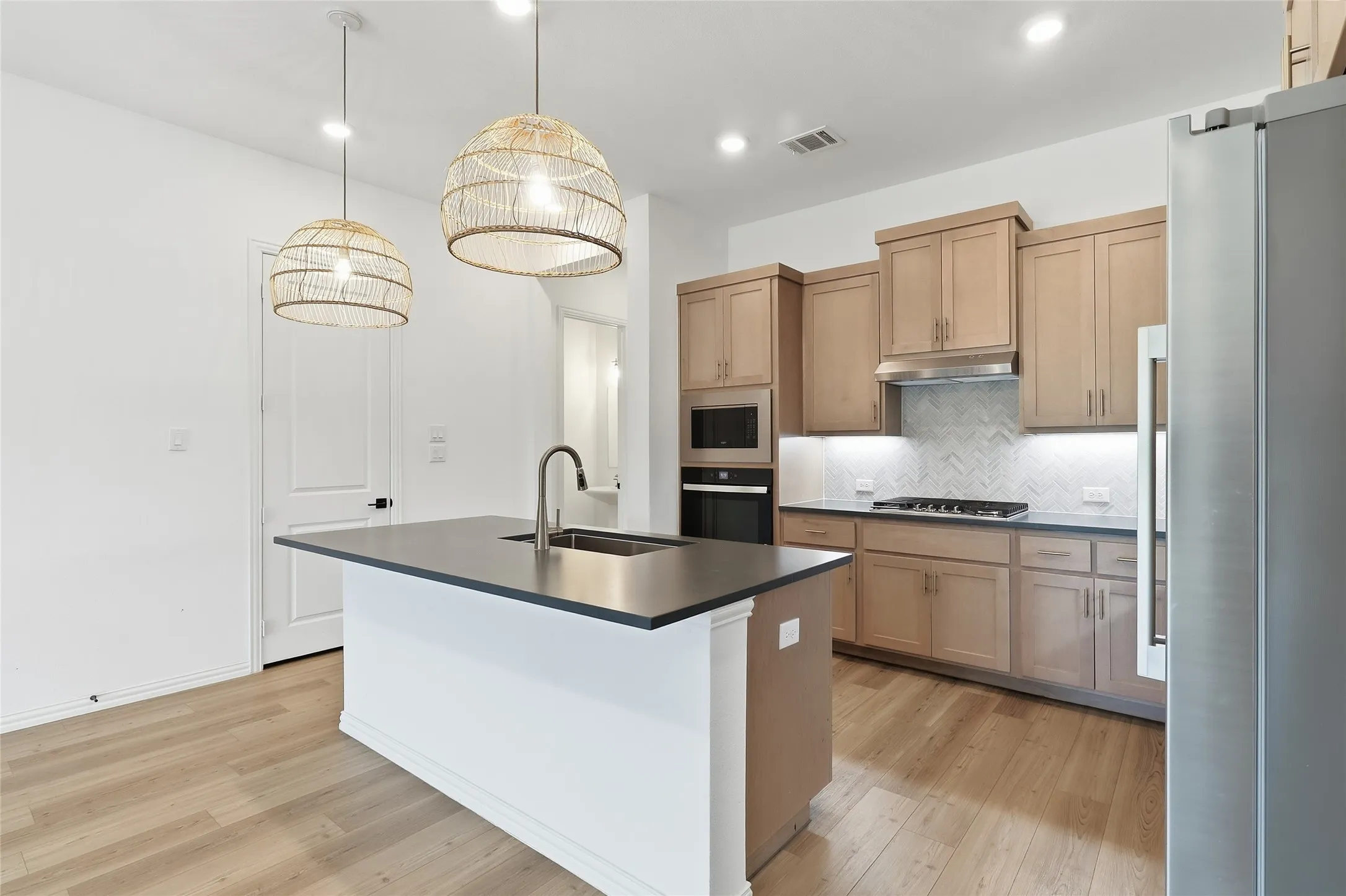 Contemporary kitchen featuring a center island with an undermount sink, pendant lighting, wood-finish flooring, and light-toned cabinetry