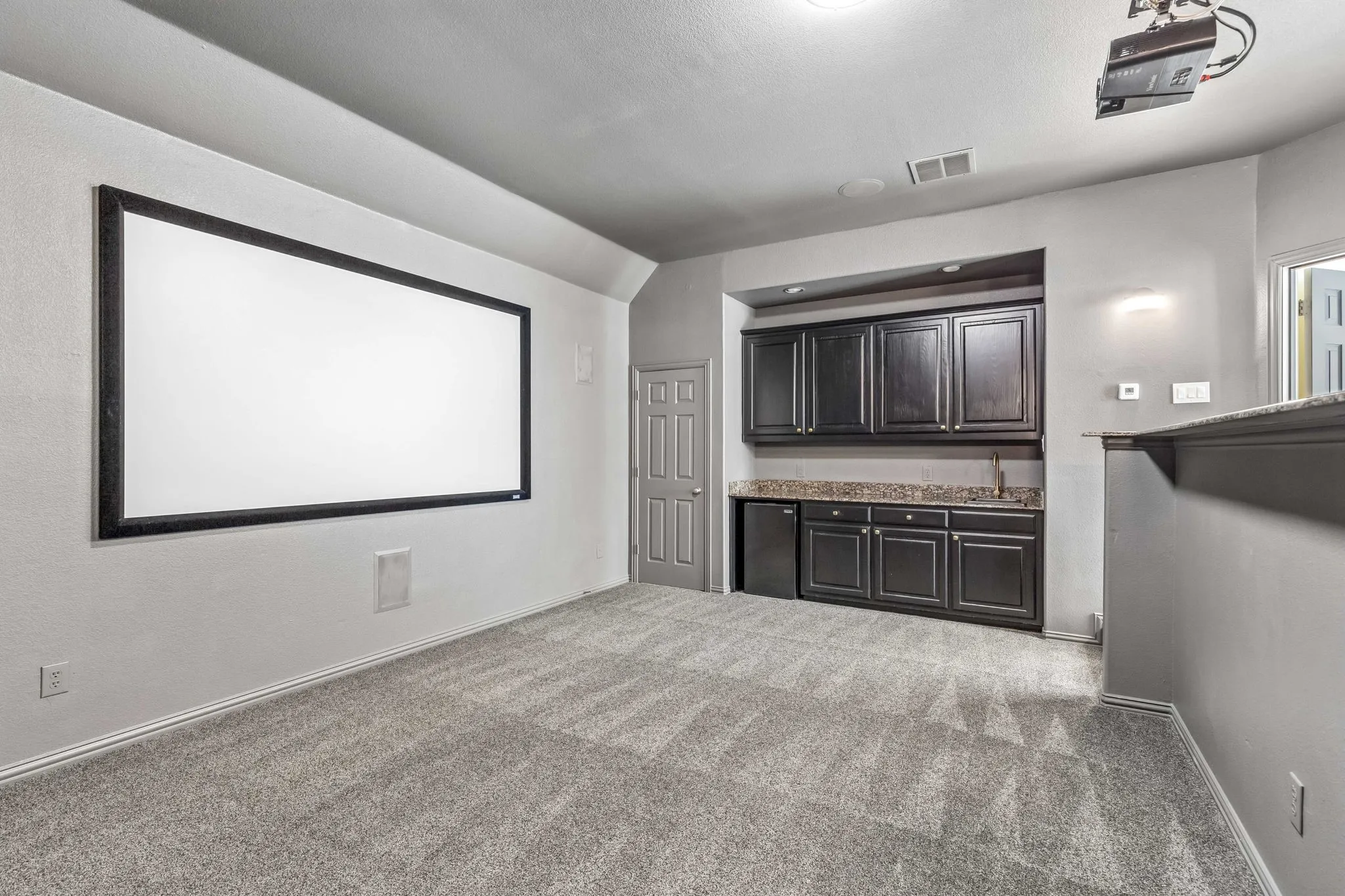 Wall-mounted projection screen, integrated wet bar with dark cabinetry, compact beverage refrigerator, recessed lighting, and built-in shelving