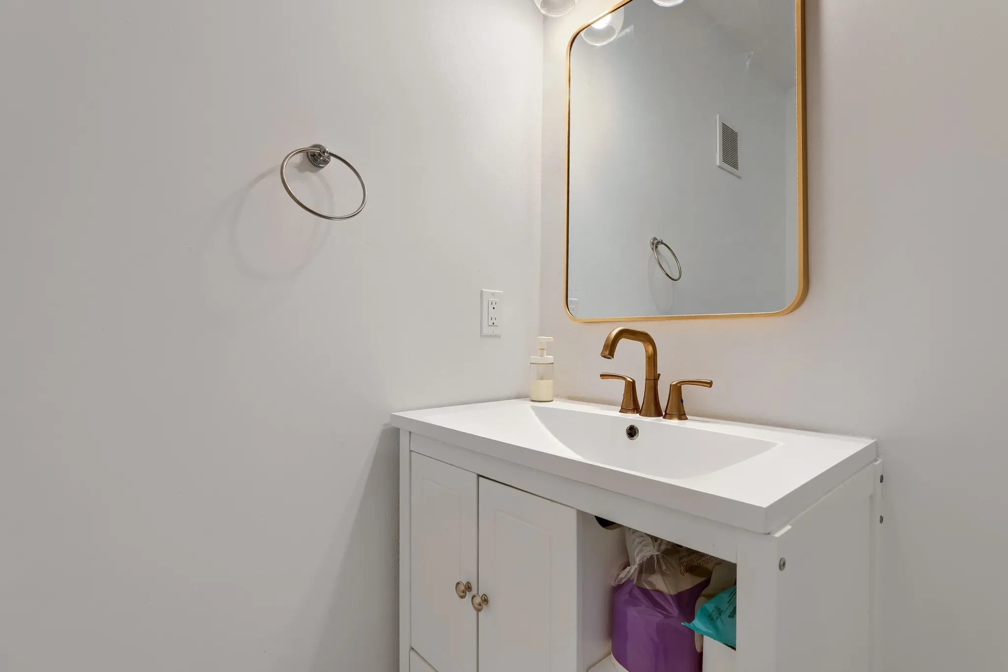Half bath, White vanity with integrated sink and brushed brass faucet, complemented by a gold-framed mirror and round wall-mounted towel holder