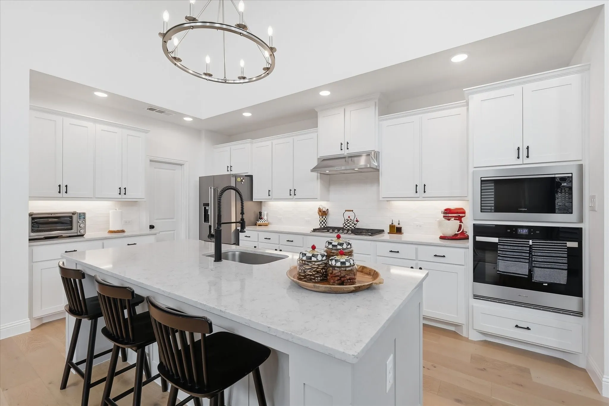 Open-concept kitchen featuring an oversized island with seating, stainless steel appliances, white shaker cabinetry with dark hardware, a single basin sink with a matte black faucet, and wood-finish flooring