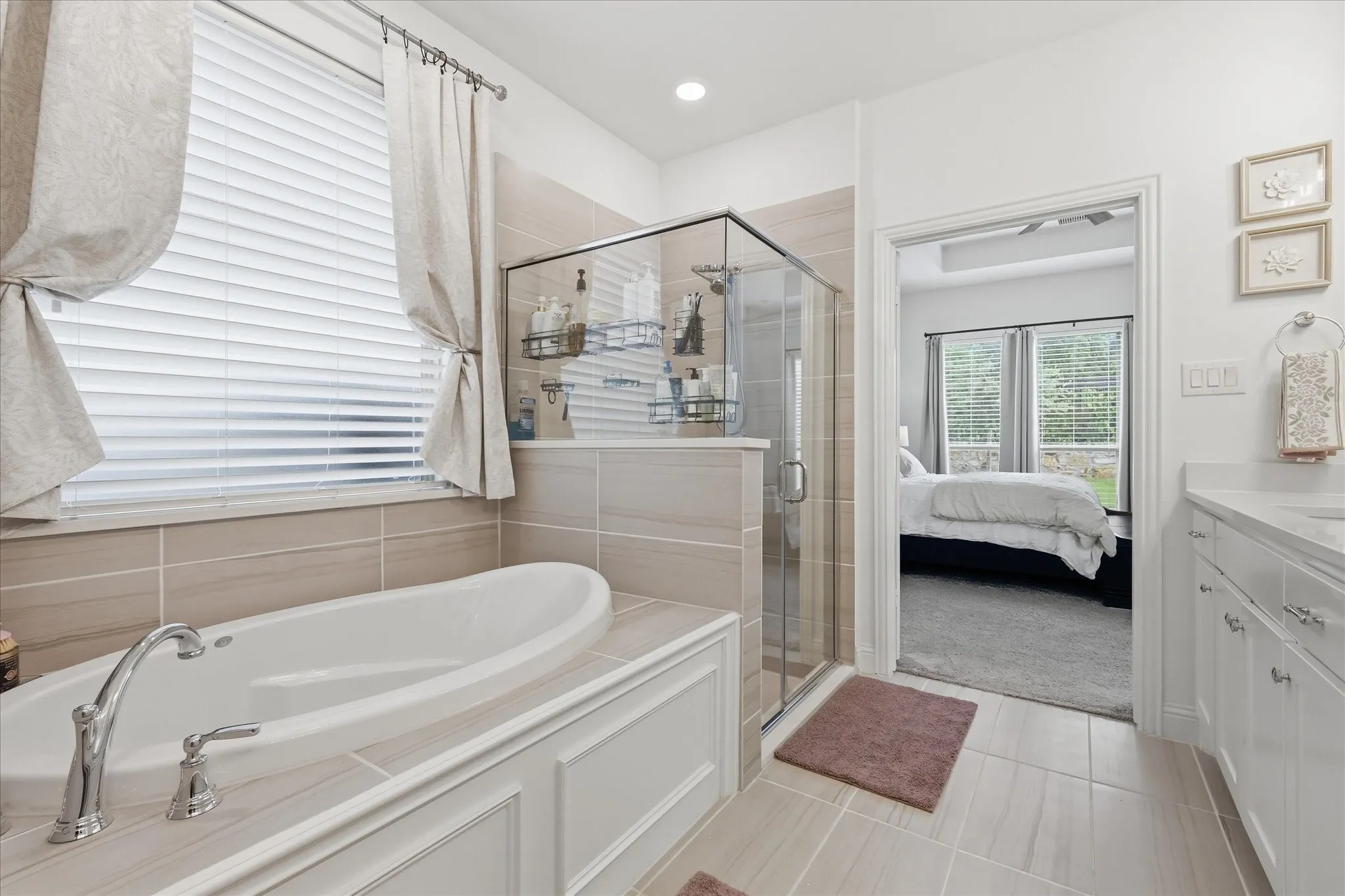 Bathroom featuring a built-in soaking tub with chrome fixtures, a glass-enclosed shower, light-toned tile flooring, and a window with blinds and drapes