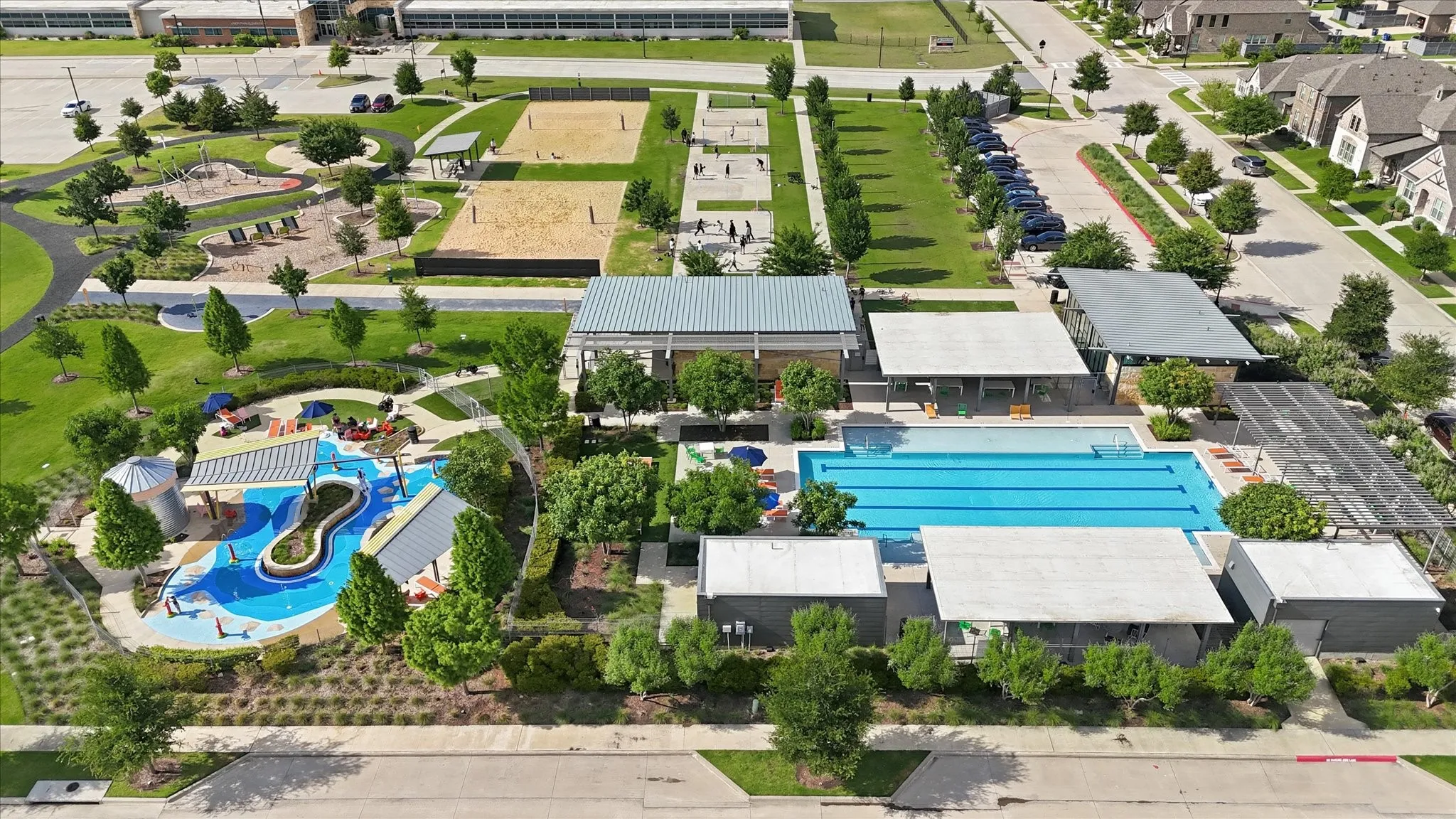 Community recreation area featuring a large swimming pool, a separate children's splash pad with shade structures, multiple sand volleyball courts, and landscaped green spaces