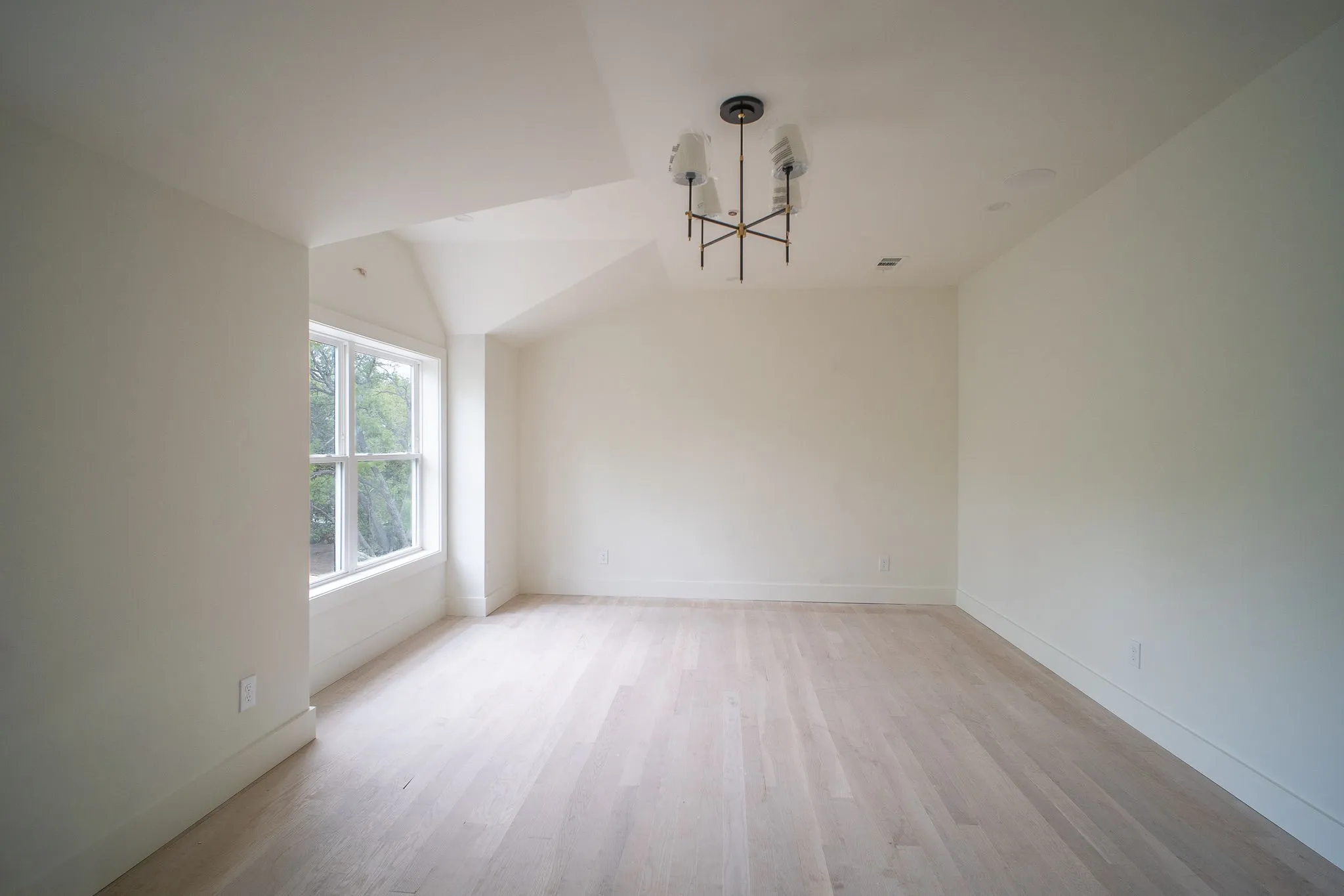 This bright room features light-colored hardwood flooring, a window providing natural light and views of greenery, and a modern ceiling light fixture