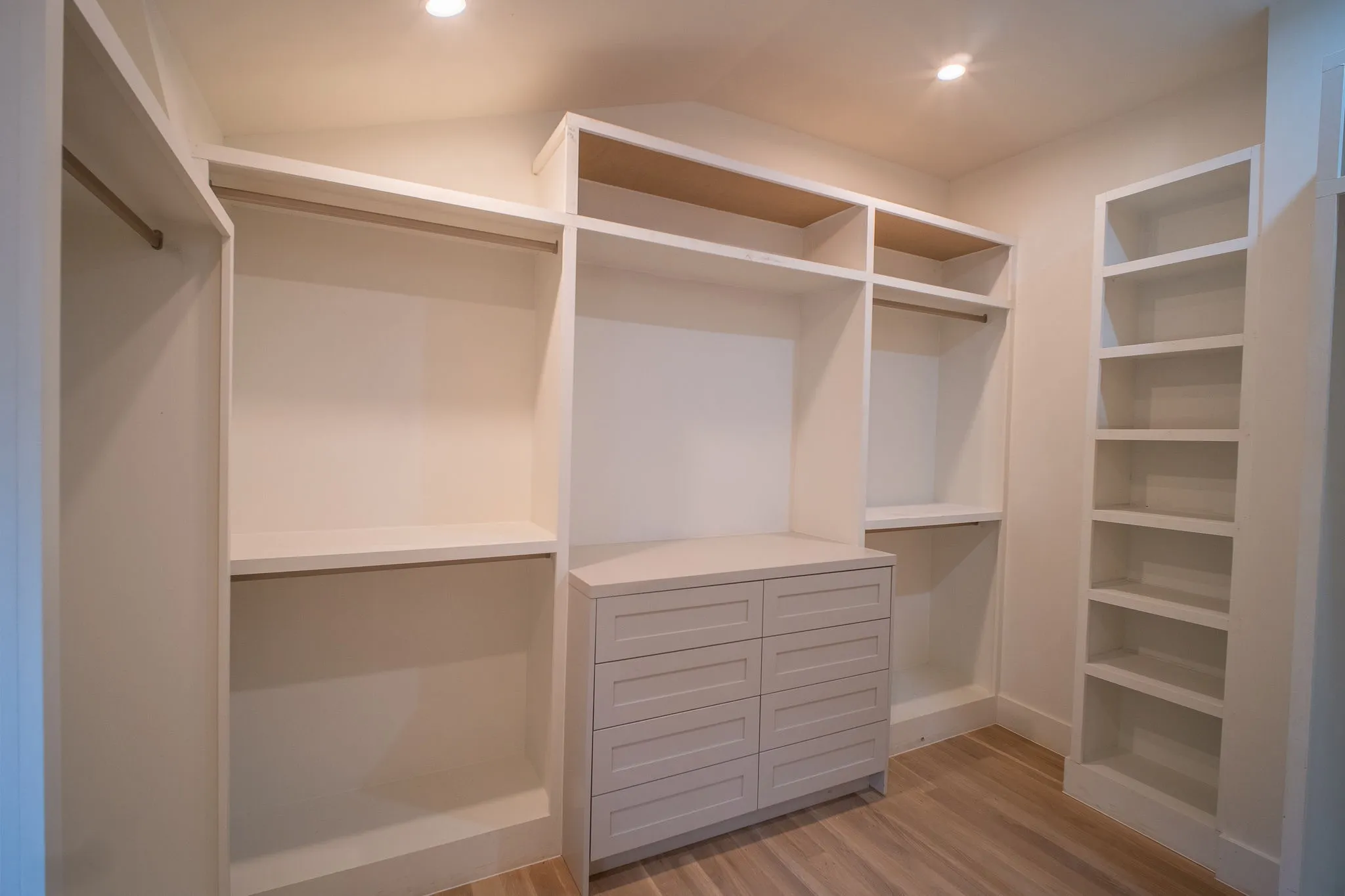 This well-appointed closet features extensive built-in shelving, hanging rods, and a central dresser with drawers, all set on light wood flooring
