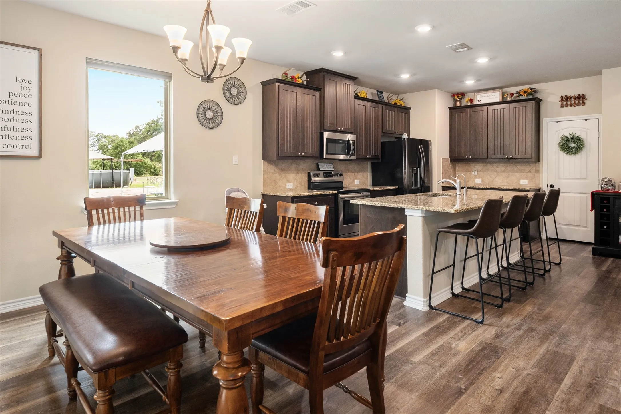 Open-concept kitchen and dining area featuring wood-finish flooring, dark wood cabinetry, a center island with countertop seating, stainless steel appliances, and a tile backsplash