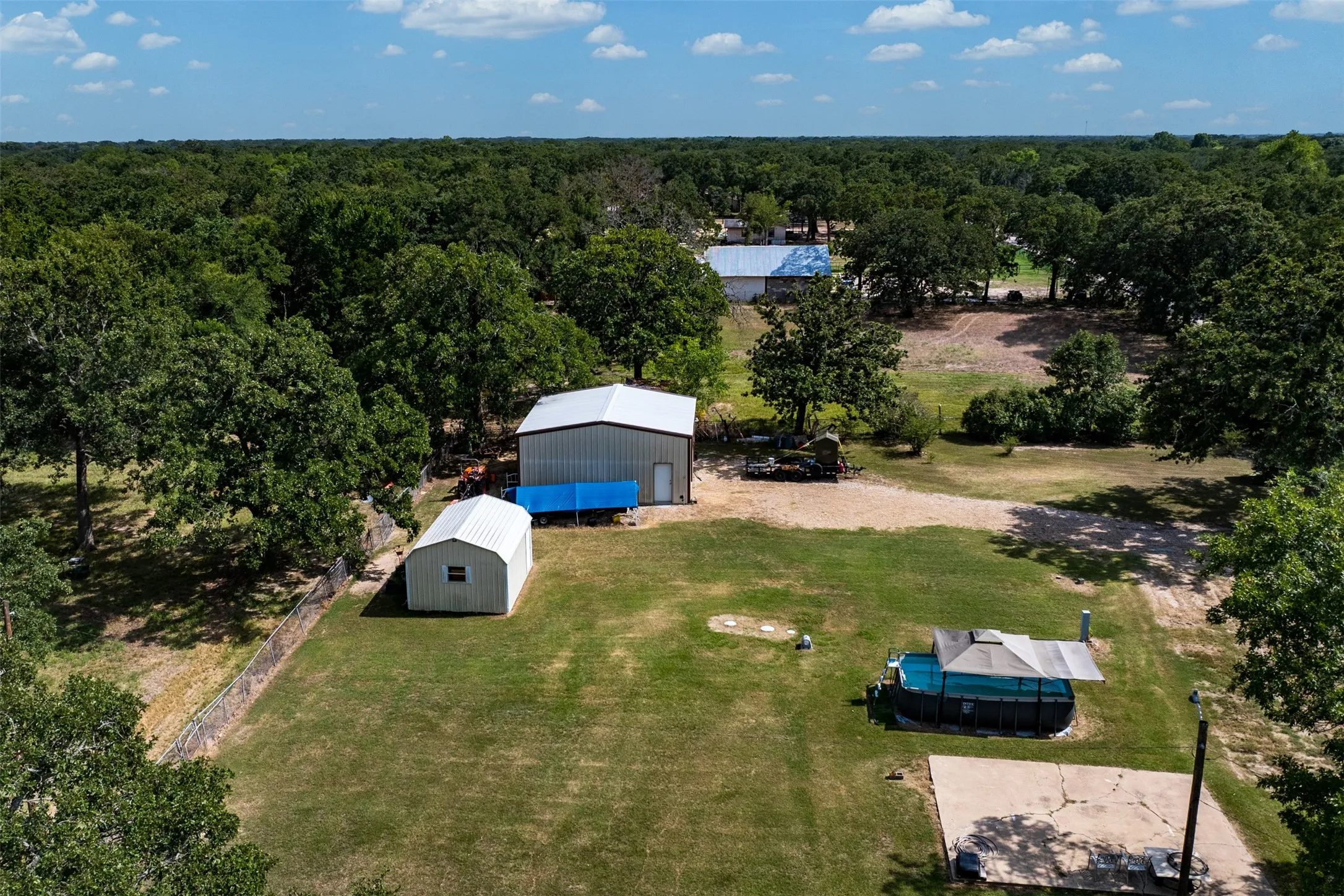 Expansive property featuring a large metal-sided outbuilding, a smaller white outbuilding, a concrete slab, and mature trees encompassing the perimeter