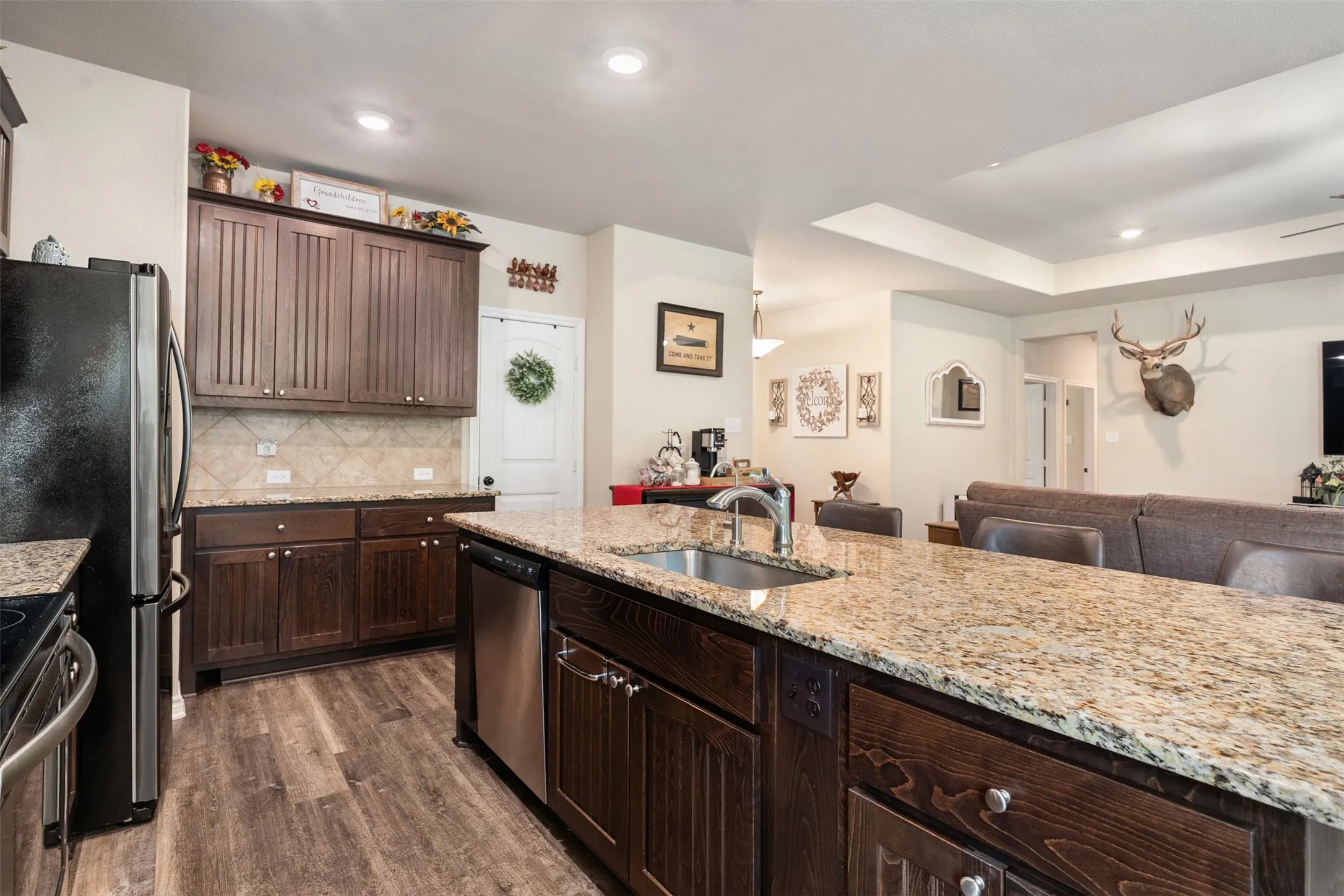 Open-concept kitchen featuring an oversized island with an undermount sink, granite countertops, wood-finish flooring, dark wood cabinetry, and stainless steel appliances