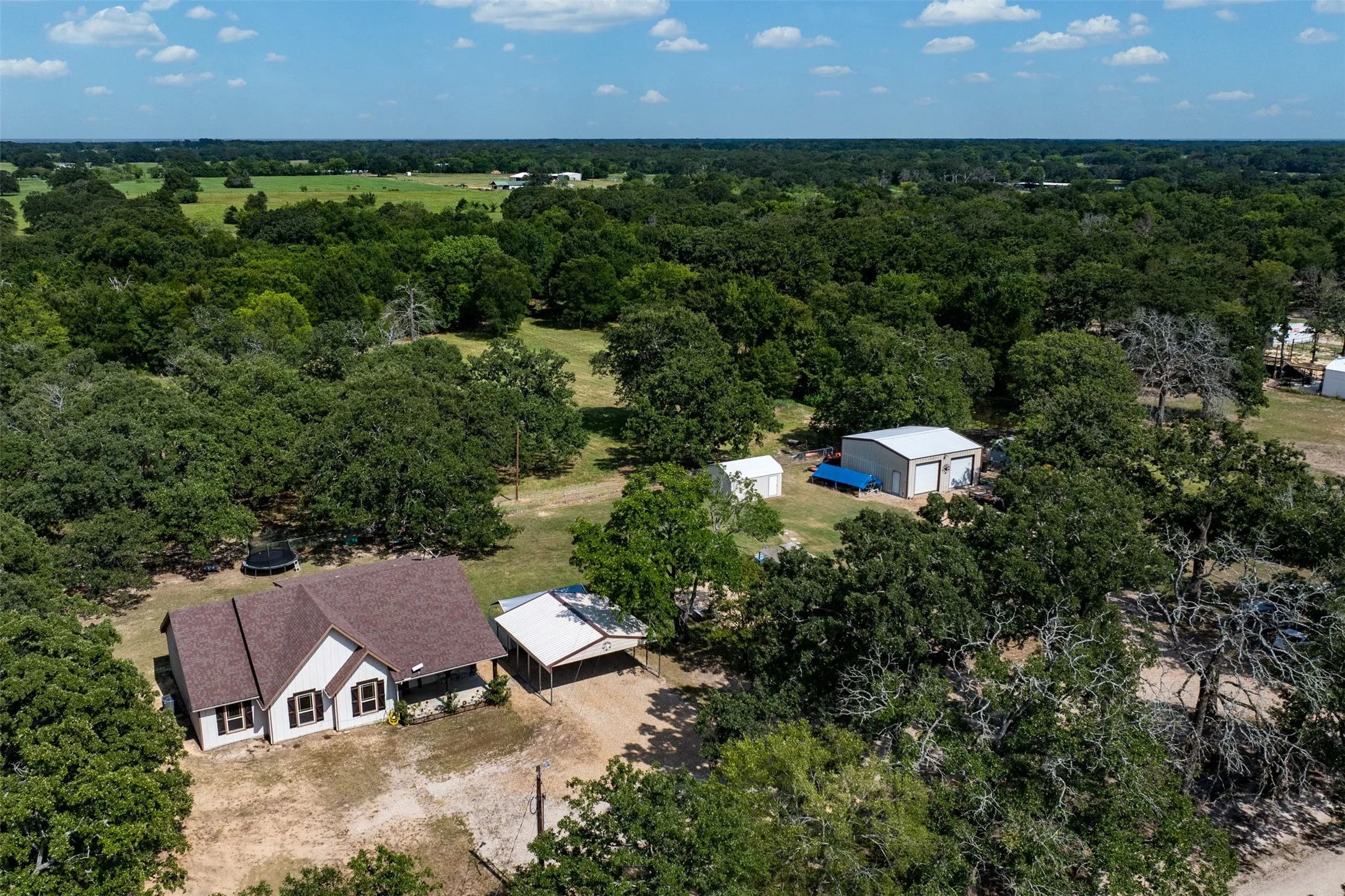 Aerial perspective showcasing a single-story residence with a brown shingle roof, accompanied by a detached metal-sided garage building with two bay doors and a white metal roof
