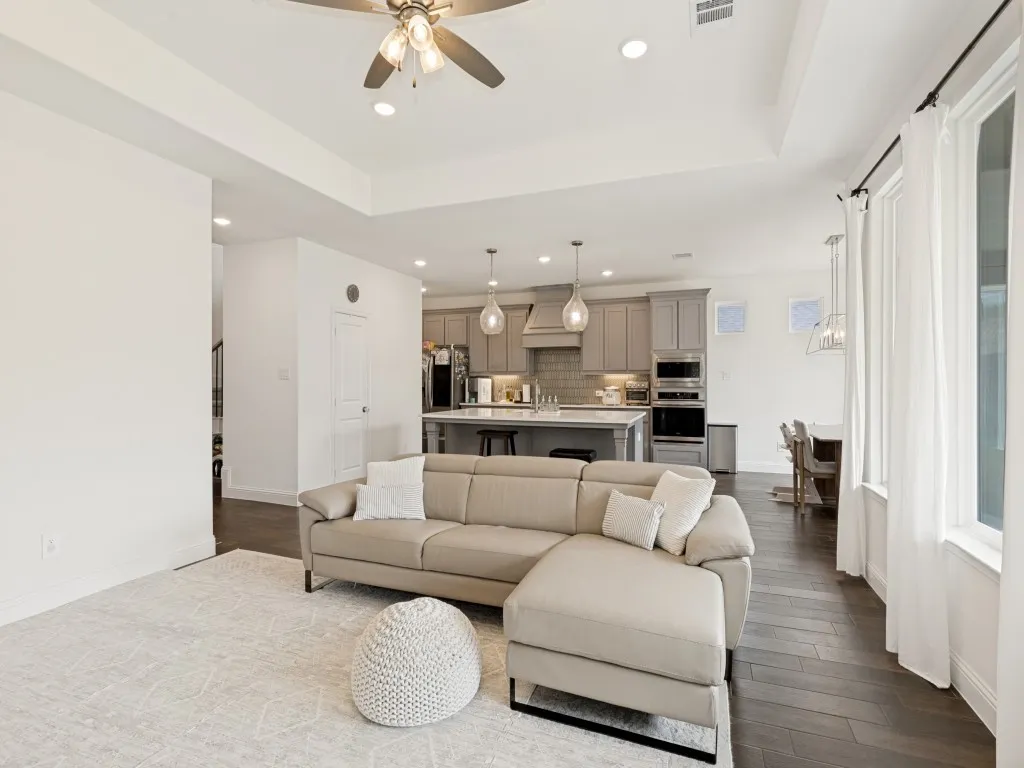 Open-concept living area featuring a tray ceiling with a ceiling fan, dark wood-finish flooring, recessed lighting, and large windows with white drapes
