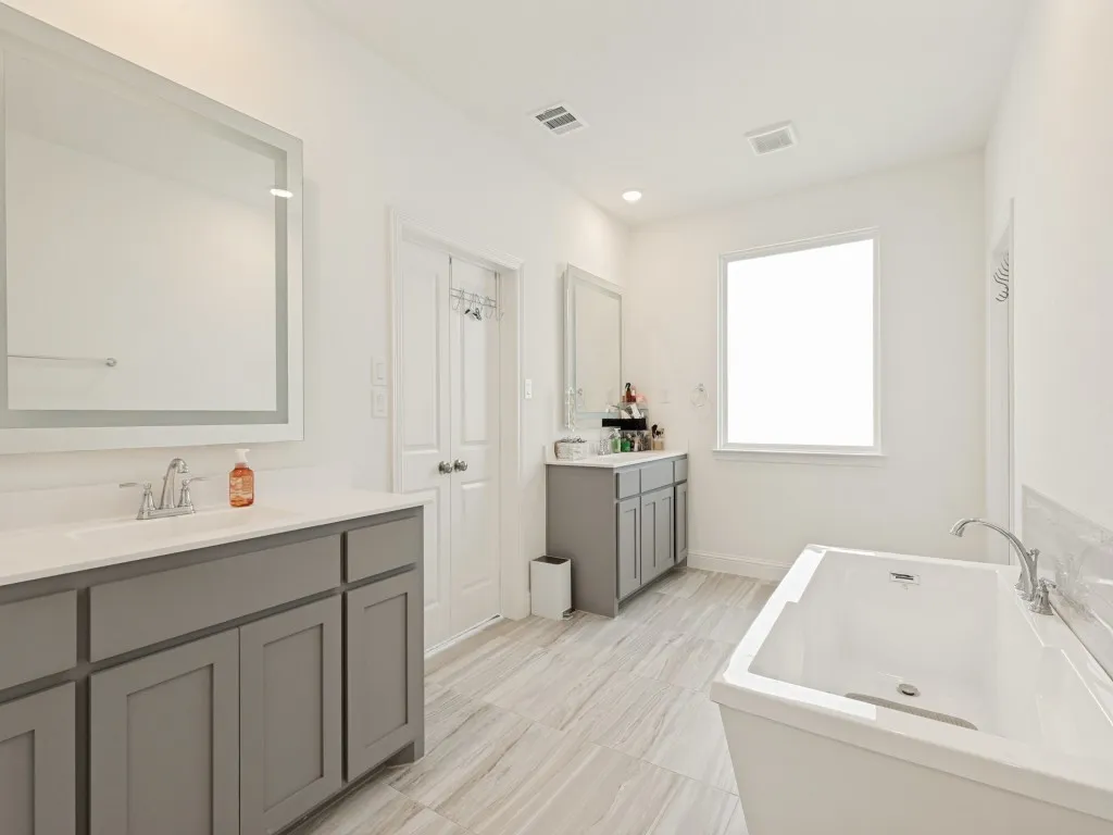 Spacious bathroom featuring a white soaking tub, two grey vanities with integrated sinks, a large window, and wood-finish flooring