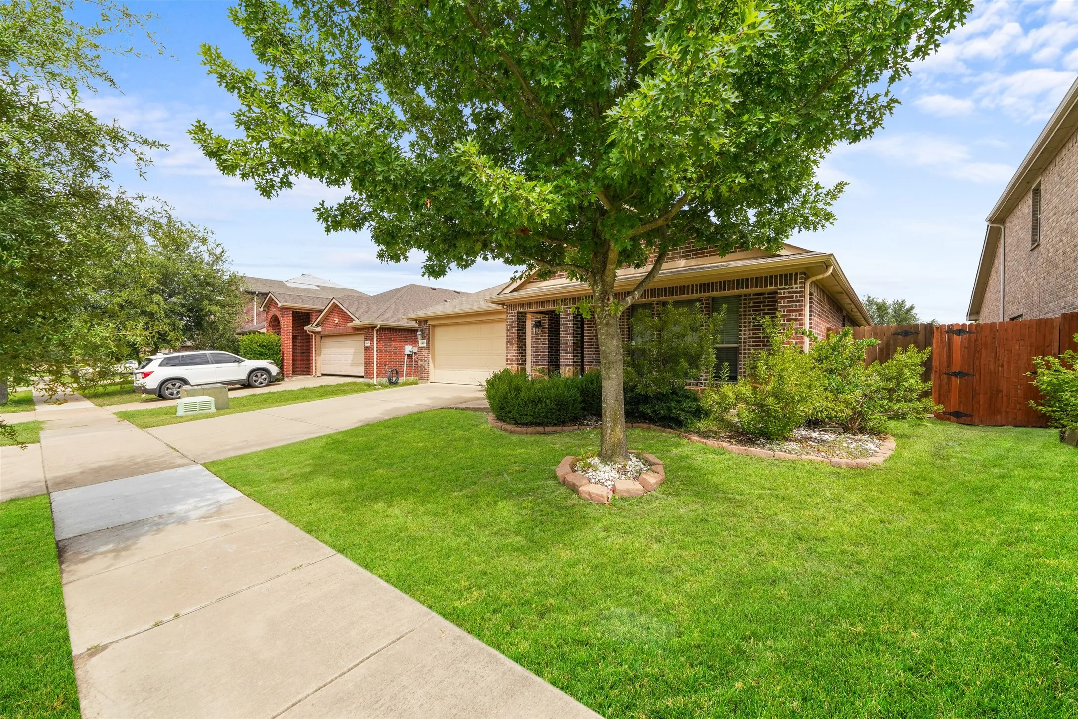 Brick-front single-story residence featuring a two-car garage, mature front yard tree, established landscaping, and a wooden privacy fence