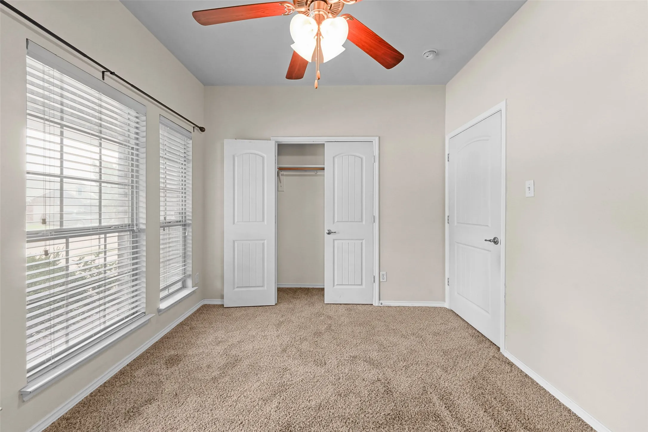 Carpeted room featuring a ceiling fan with light fixture, dual windows with horizontal blinds, and a closet with bifold doors