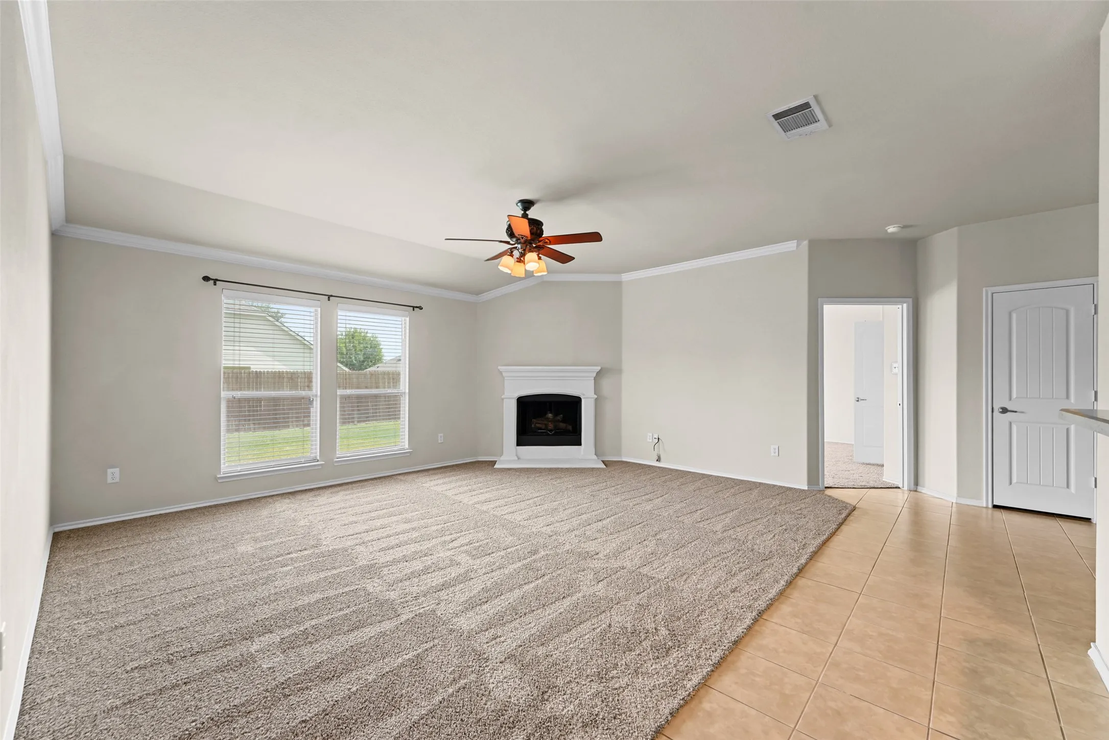 Spacious living area featuring a corner fireplace with a white mantel, ceiling fan with light fixture, large windows, and a combination of carpet and tile flooring