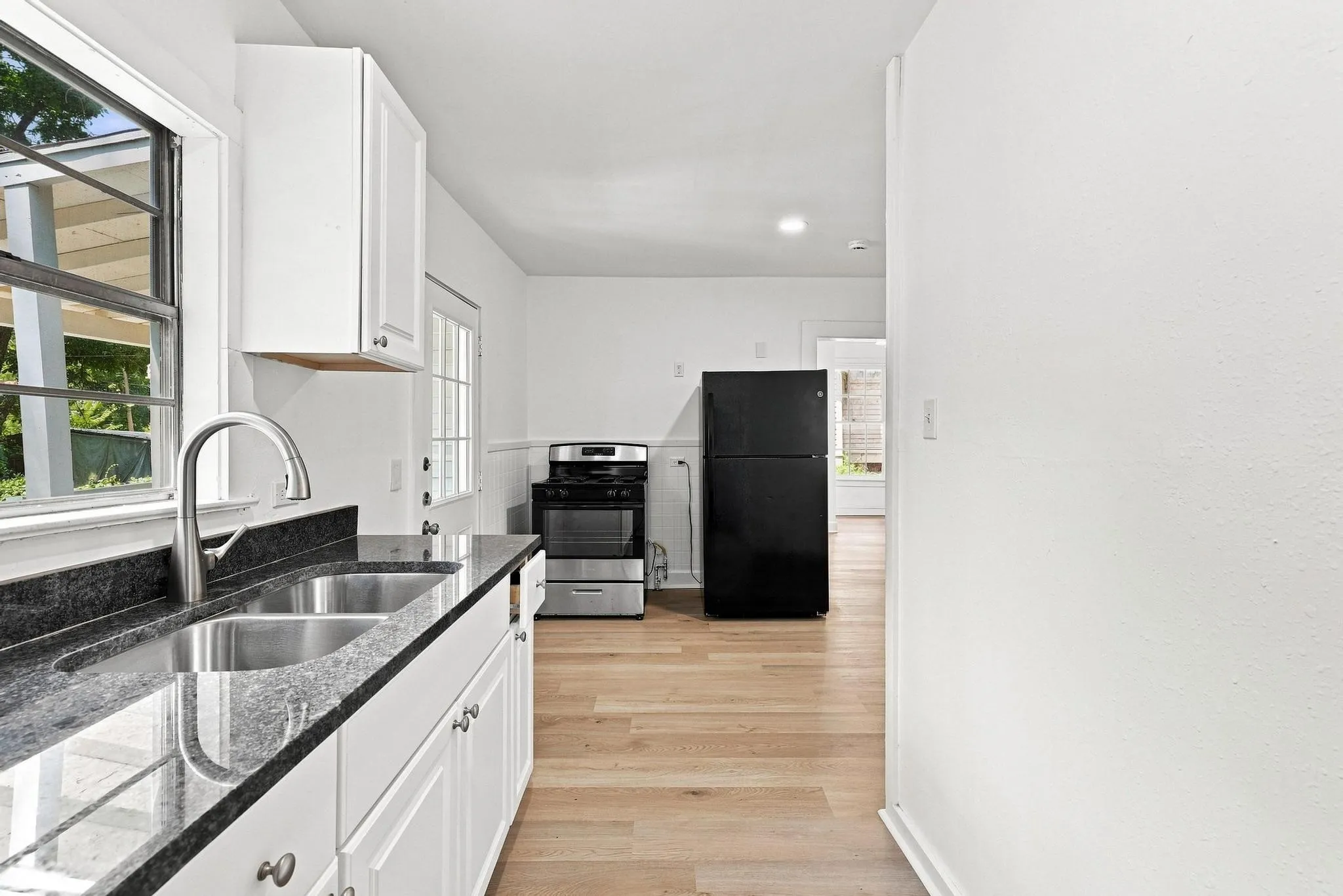 Efficient kitchen space featuring white shaker cabinetry, dark stone countertops, a dual basin stainless steel sink with a gooseneck faucet, light wood-finish flooring, and white walls