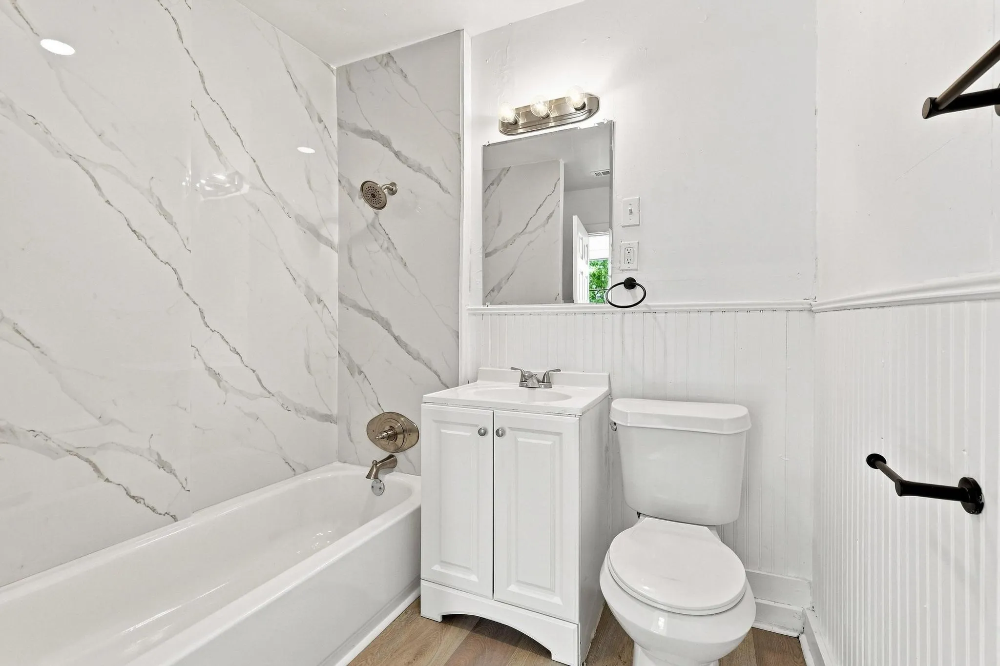 Bathroom featuring a white vanity with integrated sink, white tub with brushed nickel fixtures, marble-look shower surround, beadboard wall paneling, and wood-finish flooring