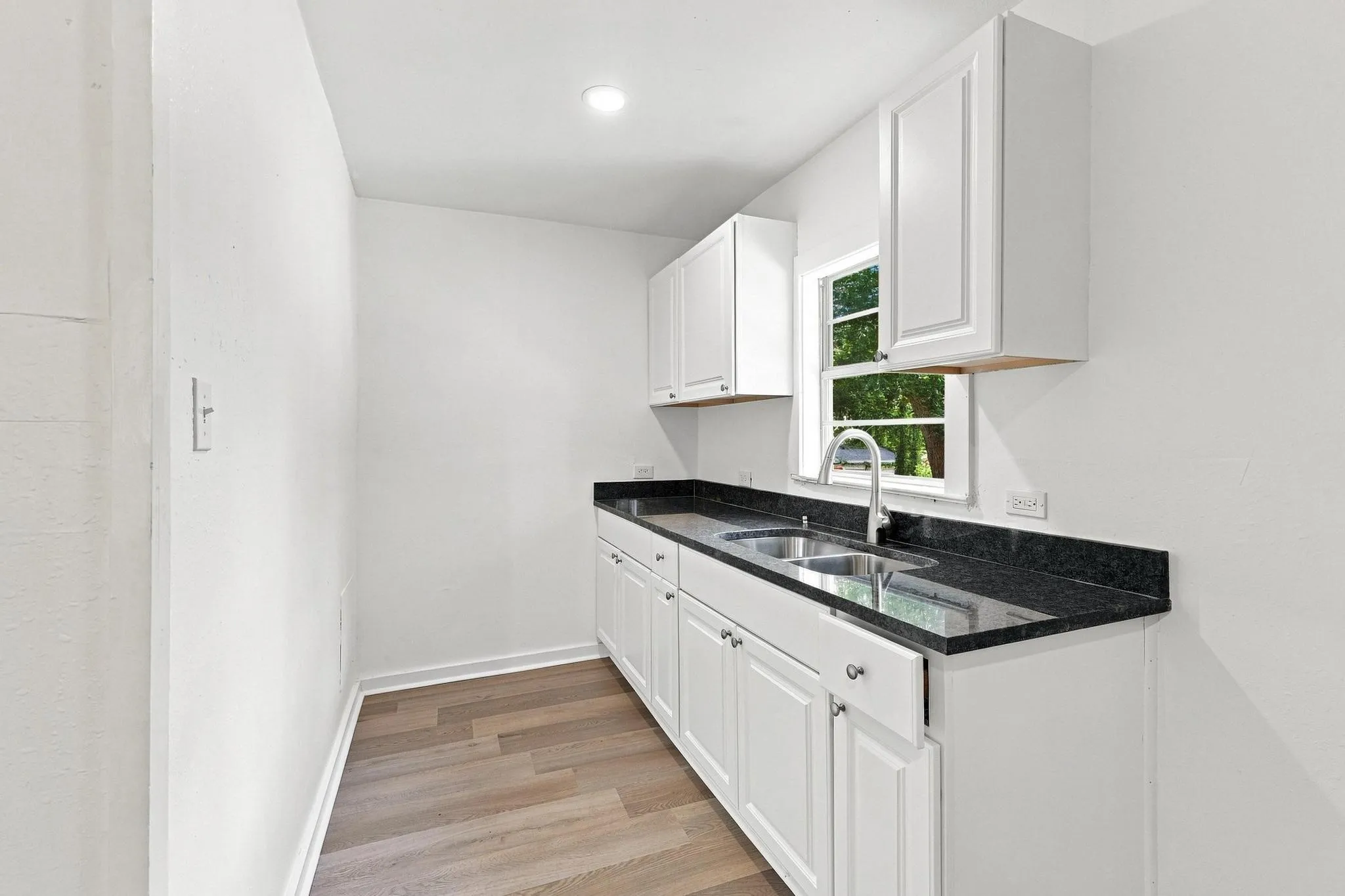 Galley kitchen featuring white cabinetry, dark stone countertops, a stainless steel double sink with a gooseneck faucet, and wood-finish flooring