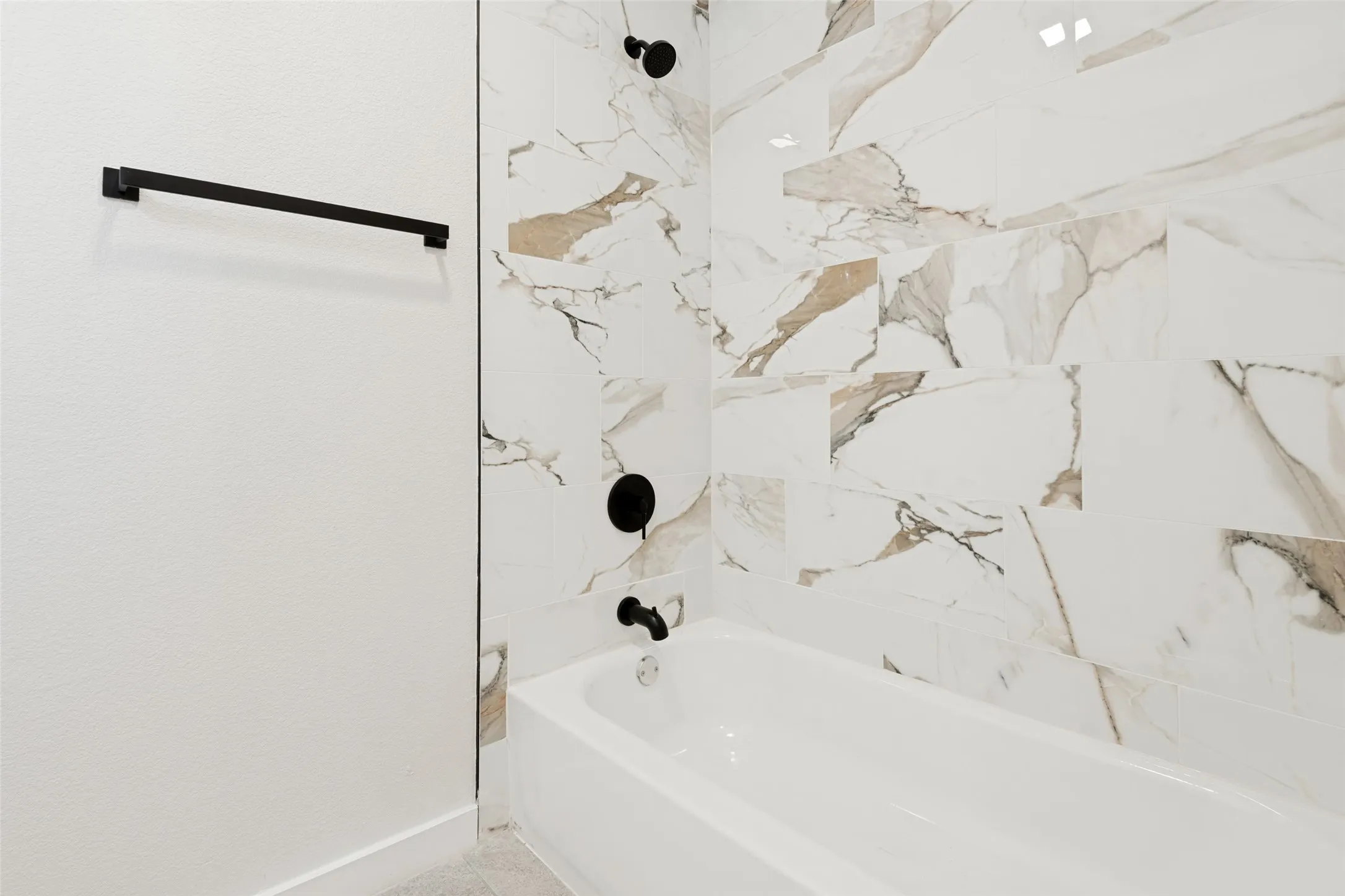 White bathtub with matte black fixtures, surrounded by large format porcelain wall tile featuring white, brown, and gray veining