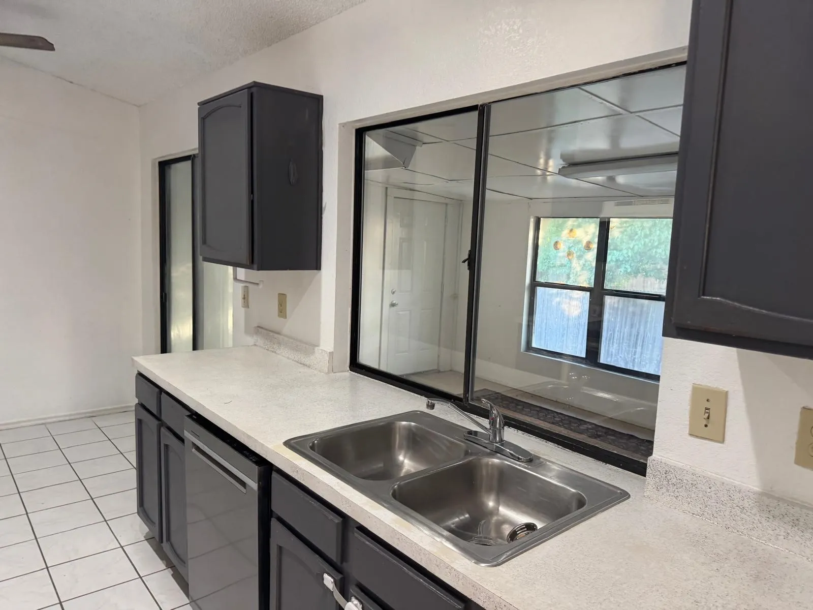 Kitchen featuring a double stainless steel sink, light-toned countertop, dark cabinetry, and white tile flooring