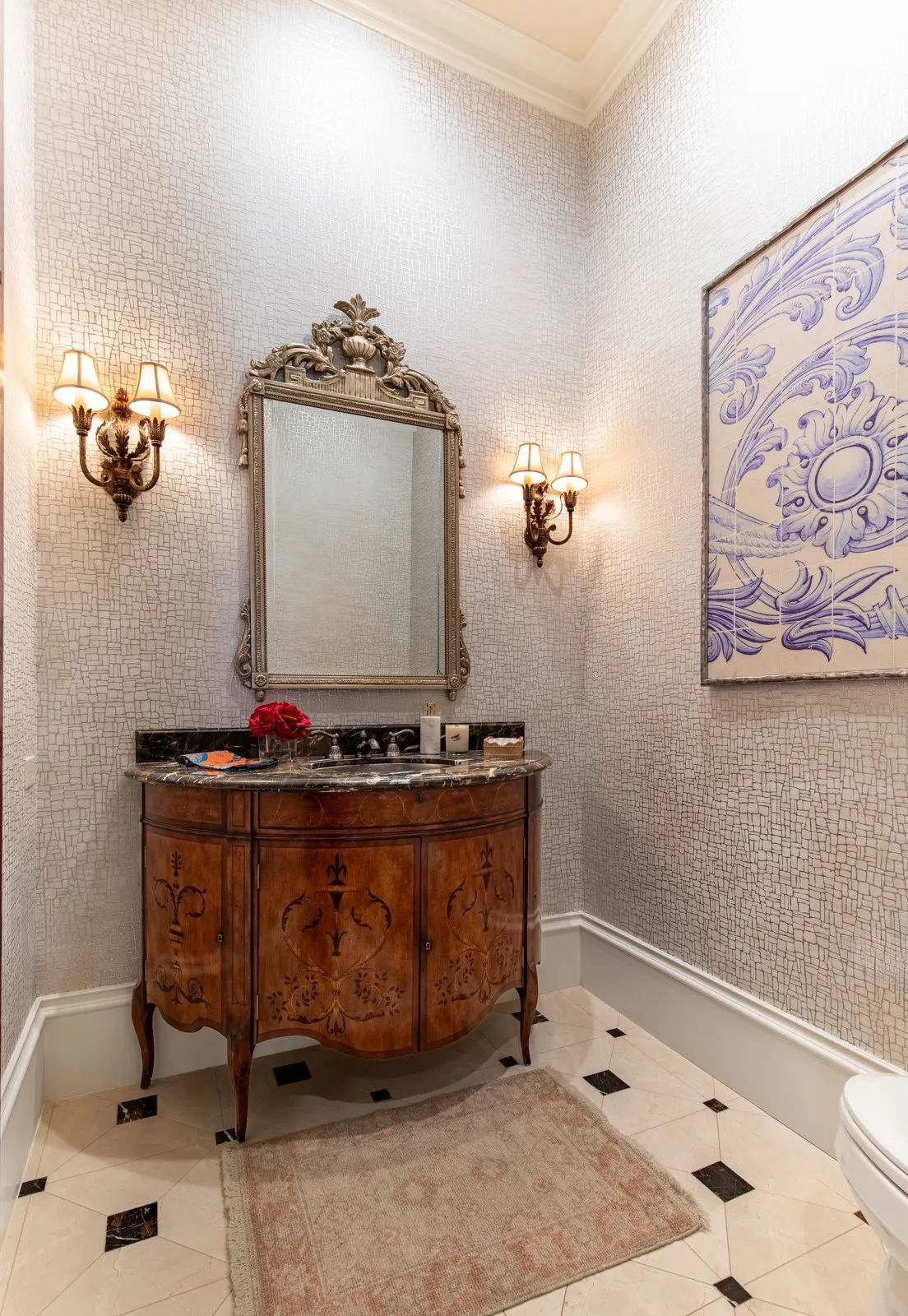 Powder room featuring an ornate wood vanity with a dark stone marble countertop, a decorative framed mirror, dual wall sconces, and stylish wallpaper