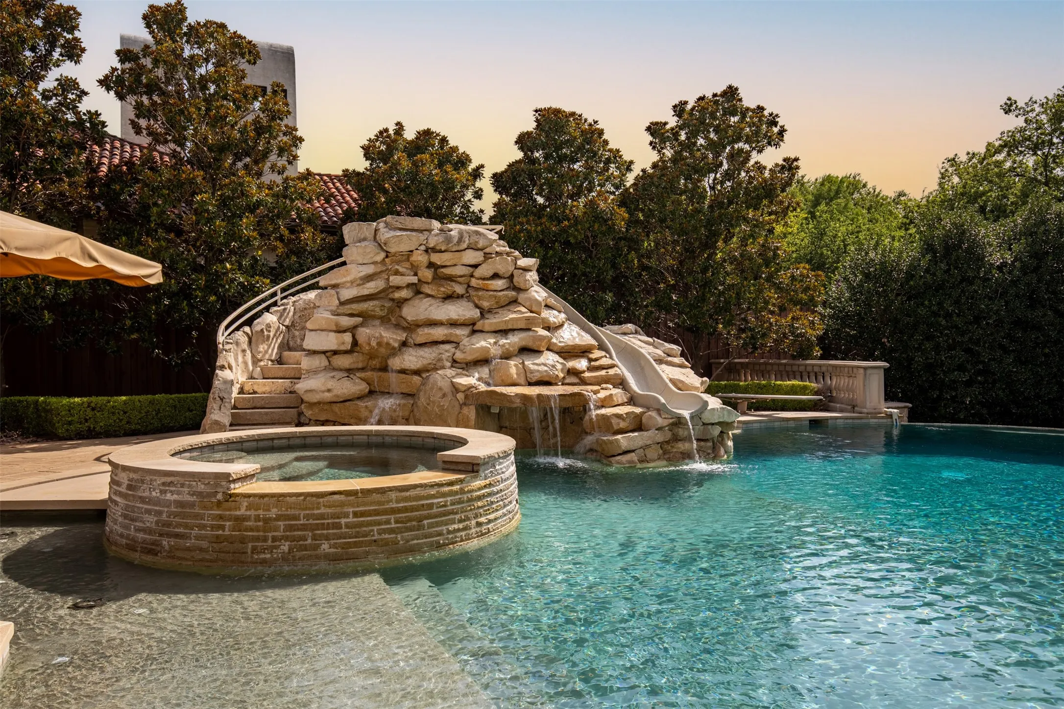 Lagoon-style swimming pool featuring a rock waterfall, integrated slide, and a raised circular spa with stone-finish exterior