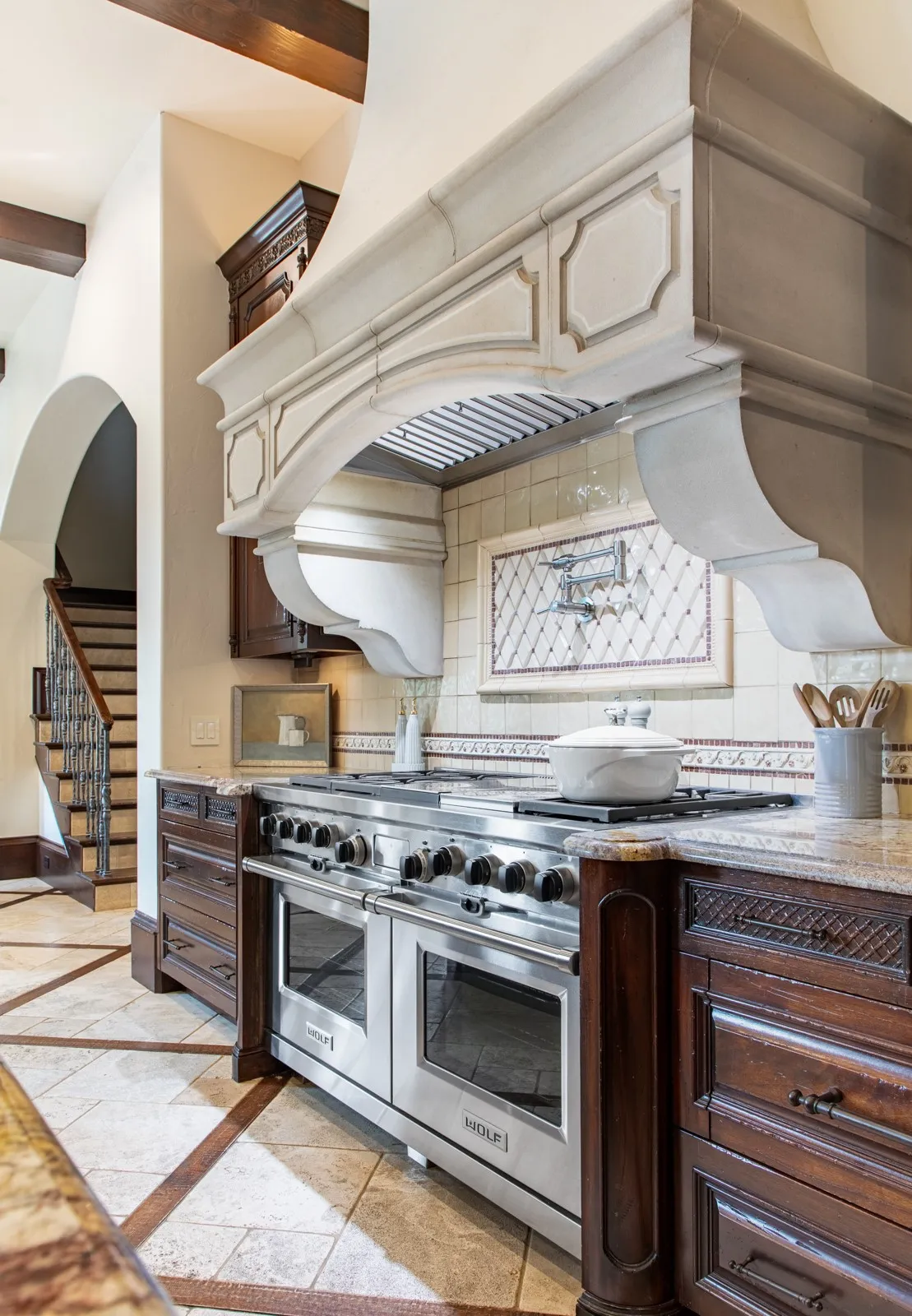 Gourmet kitchen featuring a substantial decorative range hood, professional-grade Wolf stainless steel gas range, rich wood cabinetry, granite countertops, and patterned floor tiling with wood inlay