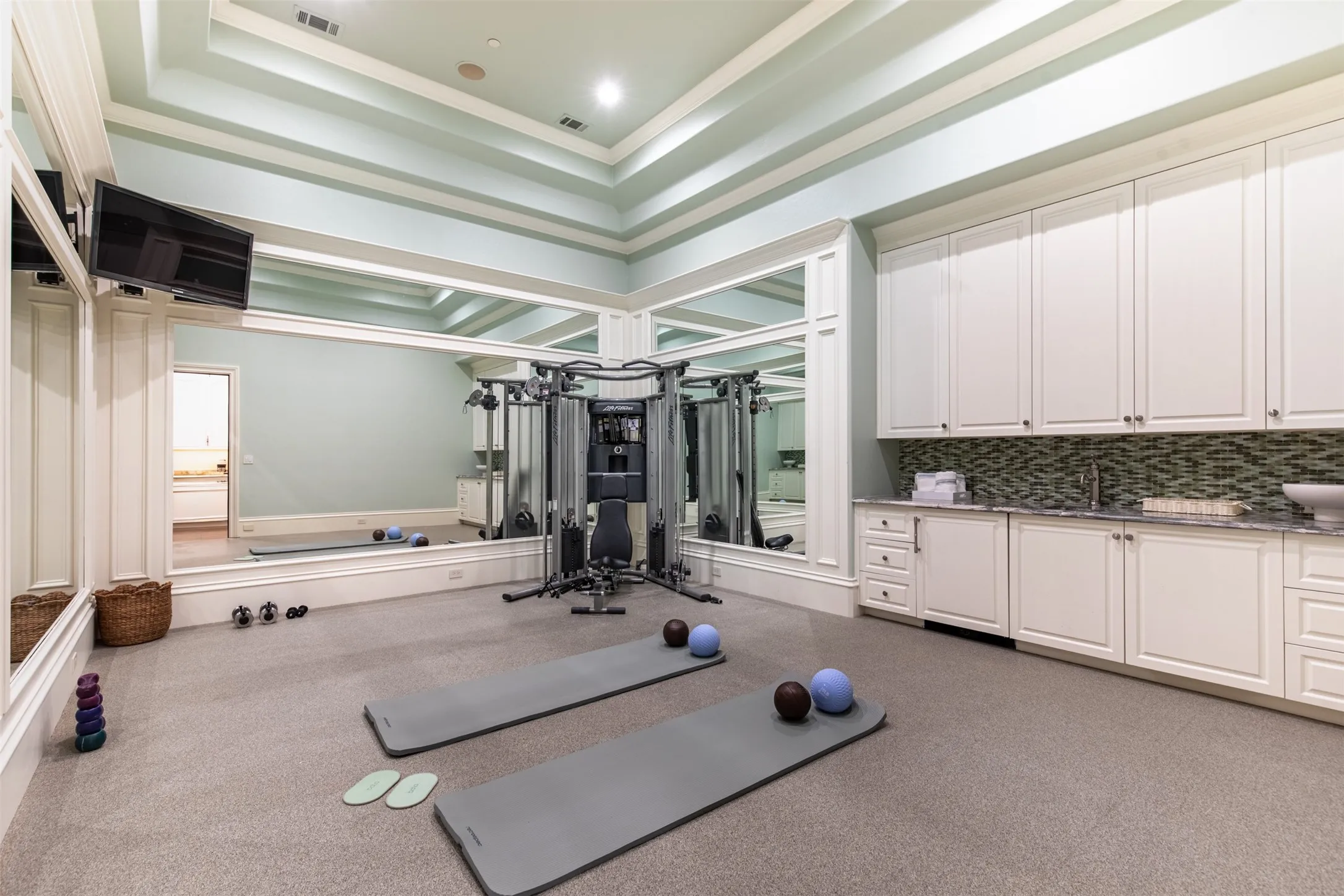 Located in primary suite area, the dedicated exercise room features a coffered ceiling, wall-mounted mirrors, built-in cabinetry with integrated wet bar, and wall-to-wall carpeting.