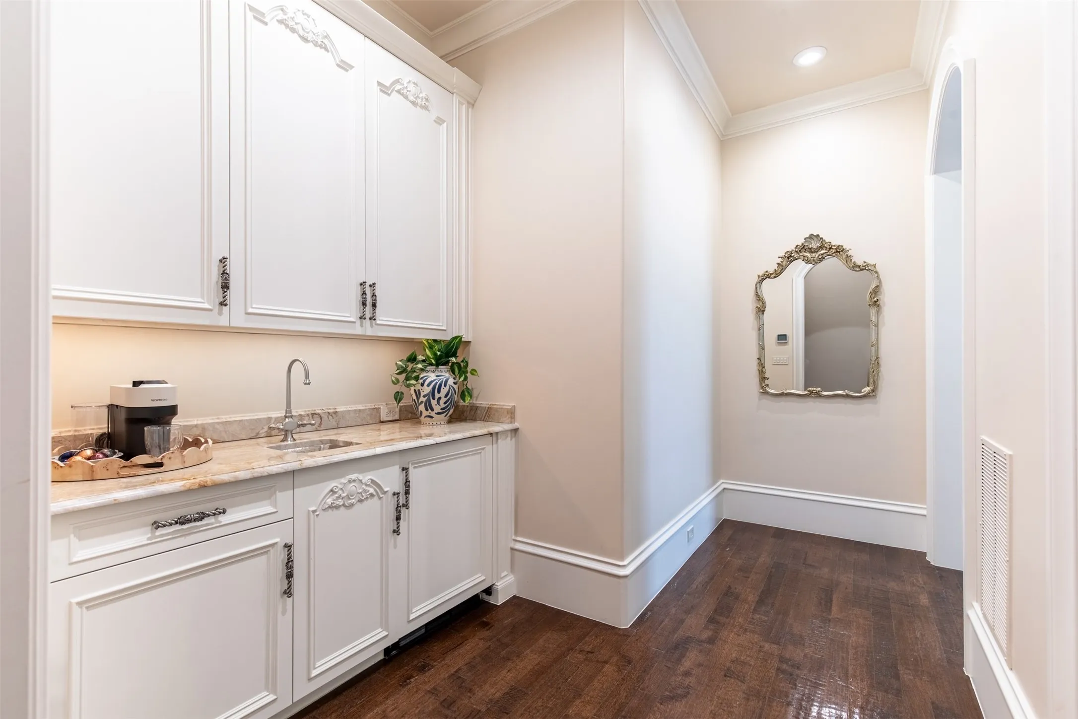 Built-in custom coffee bar with decorative molding, under-cabinet lighting, a polished stone countertop, and a single basin sink with a chrome faucet. Located upon entry to the primary bedroom suite