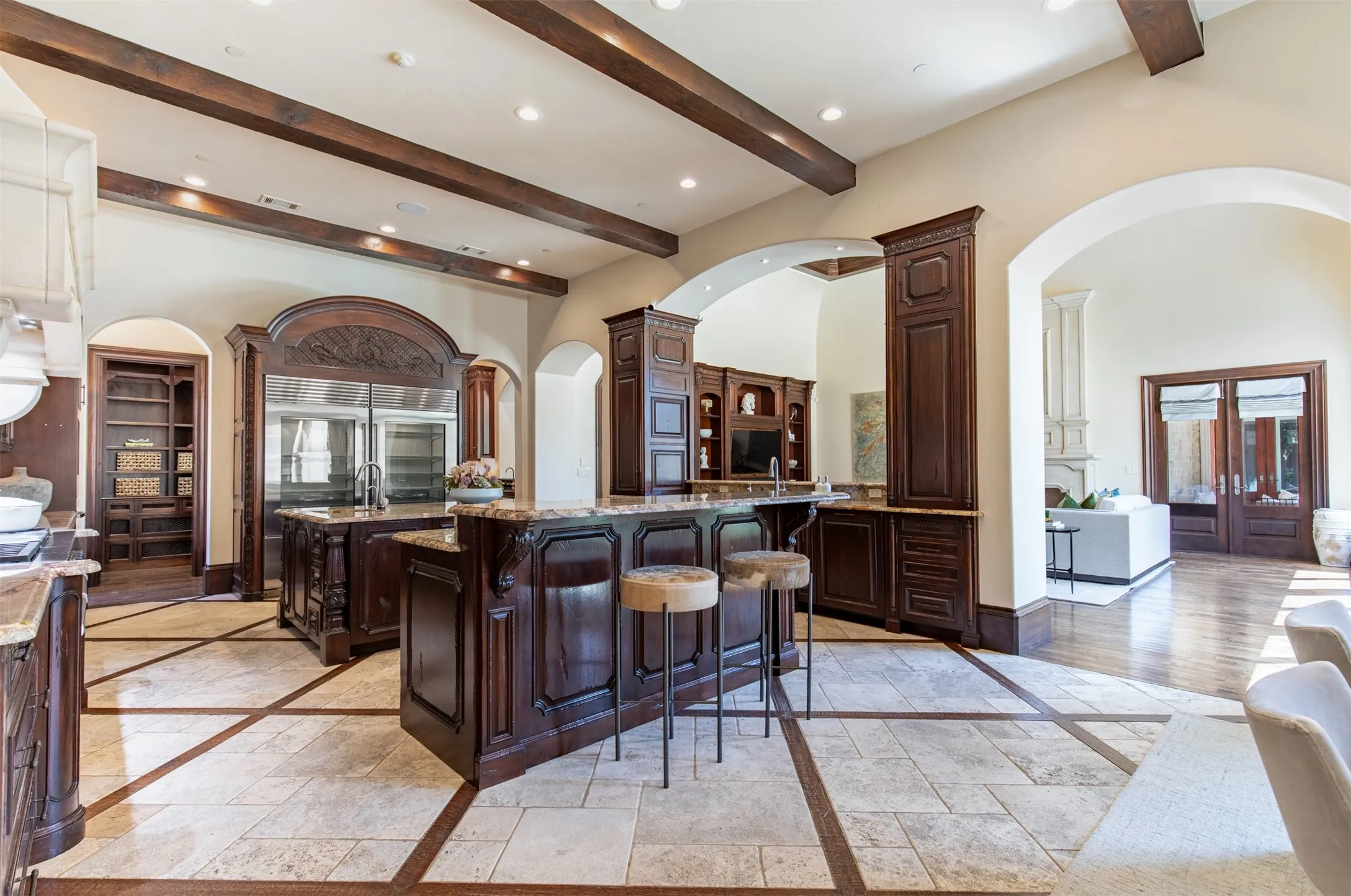 Expansive kitchen featuring a large central island with seating, dark wood cabinetry, integrated stainless steel Wolf and Sub-Zero appliances, and a coffered ceiling with exposed wood beams