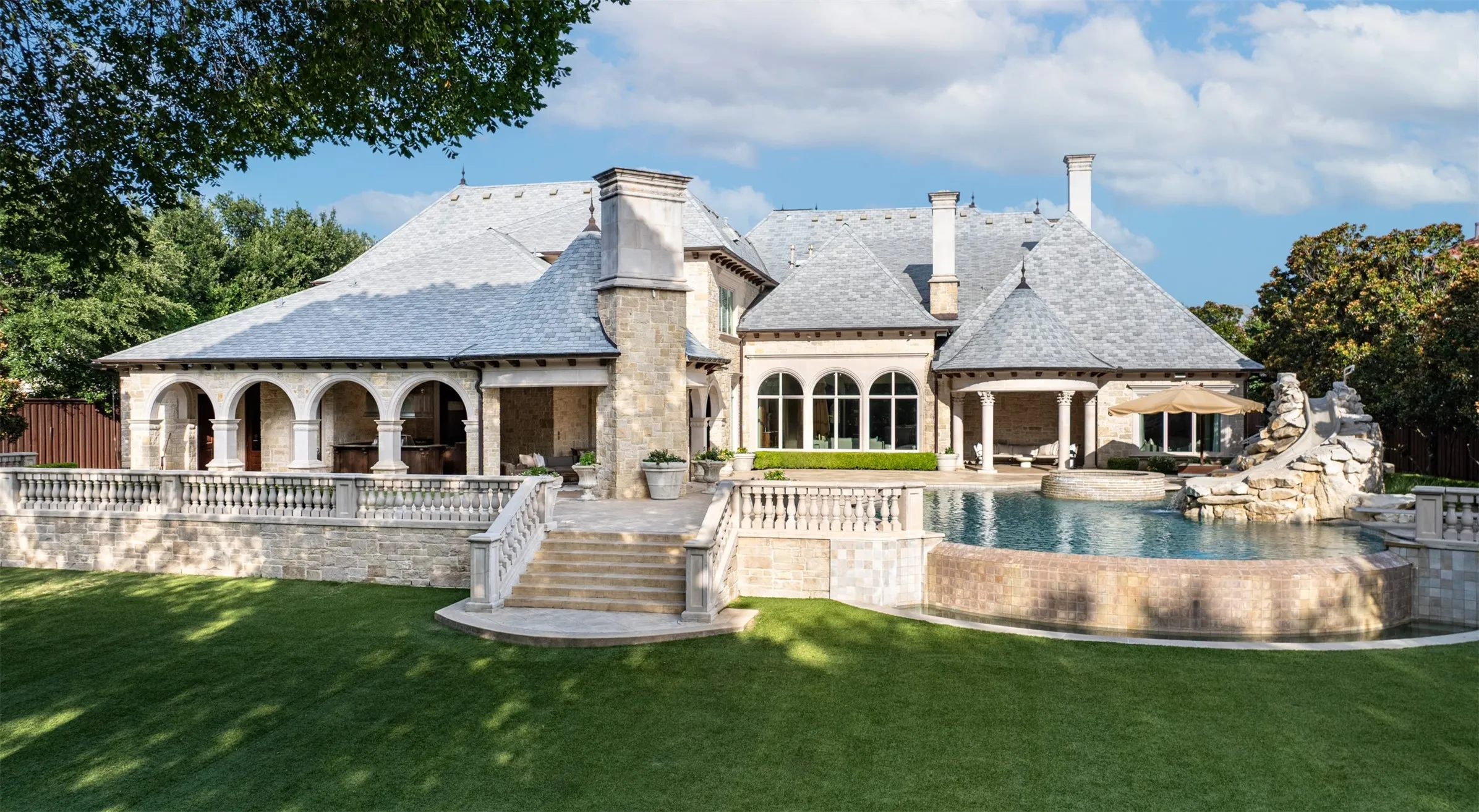Expansive rear exterior featuring a multi-tiered stone patio with balustrade, a large swimming pool with integrated spa, and a natural stone waterfall feature
