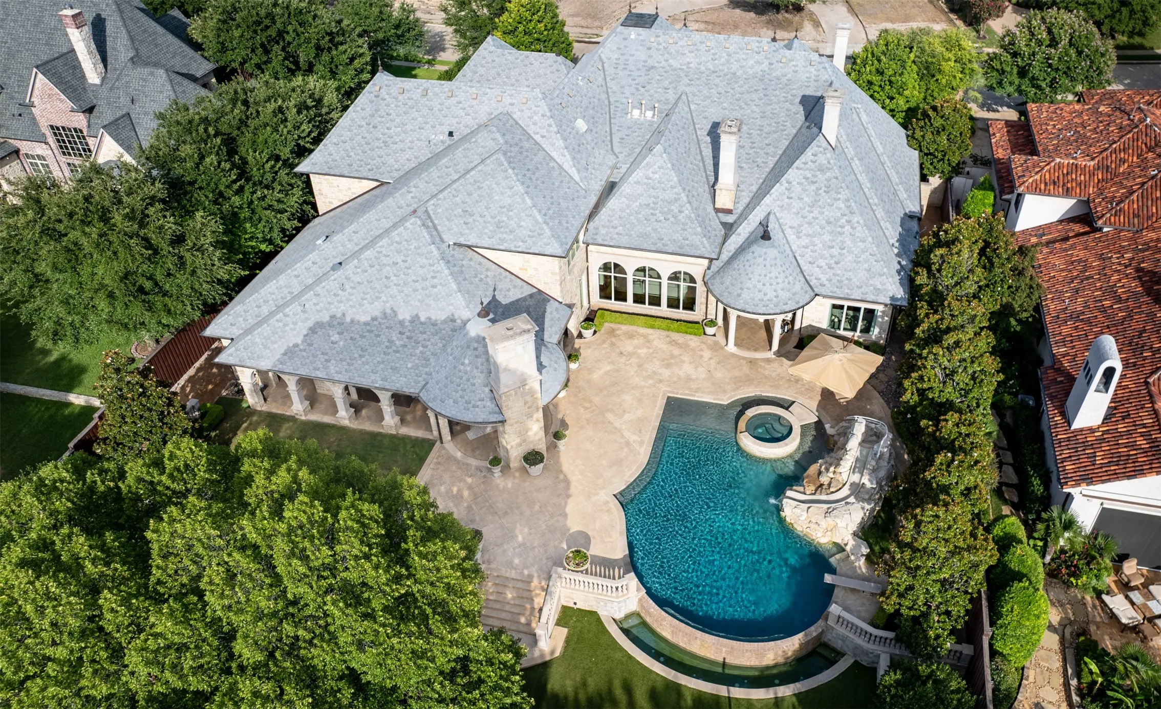 Spacious exterior featuring a multi-gabled roof, stone façade, a large free-form pool with integrated spa and rock feature, extensive hardscaping, and mature trees