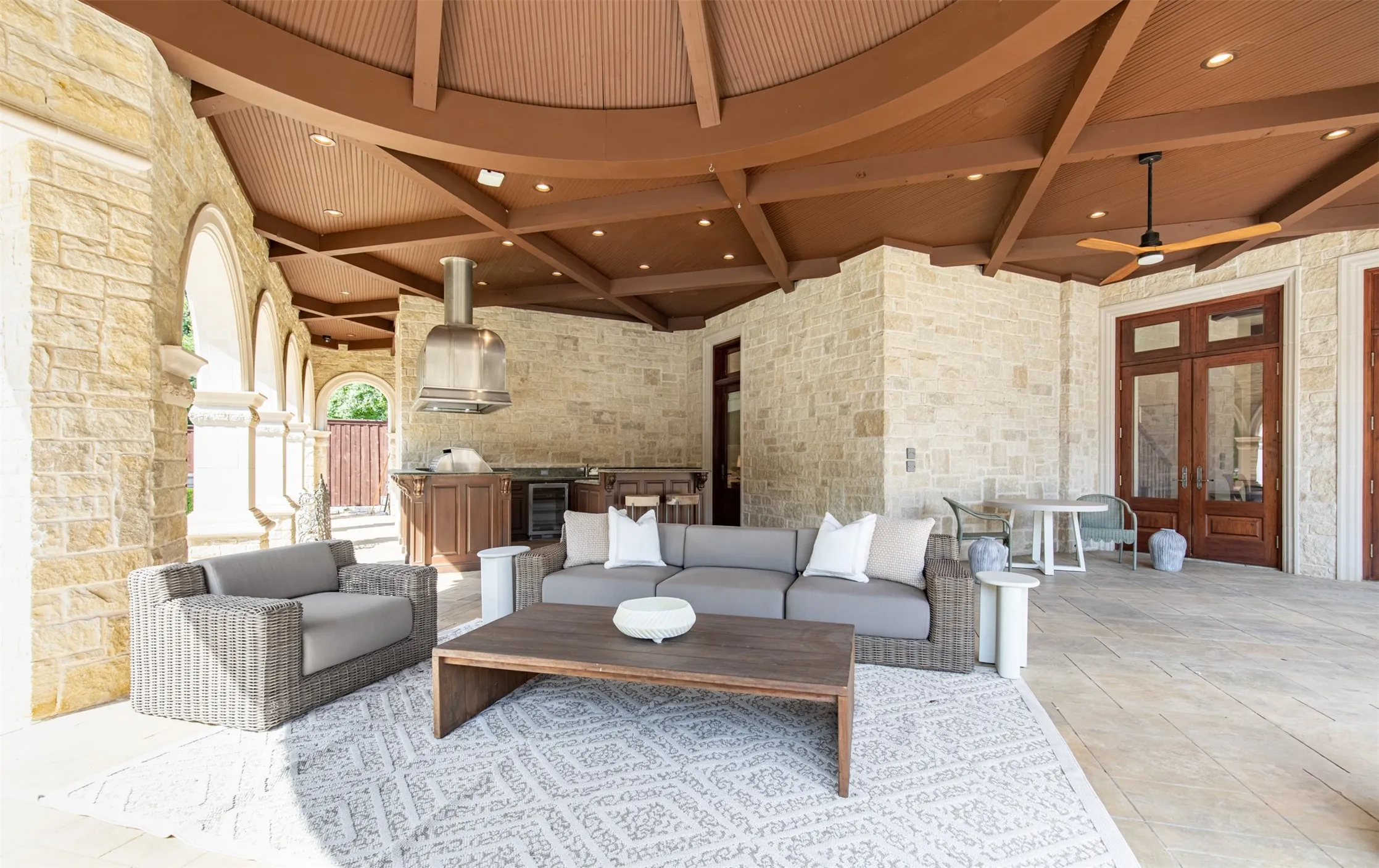 Expansive outdoor lanai featuring a full outdoor kitchen with stainless steel range hood, stone facade exterior, a wood-paneled ceiling with recessed lighting, and multiple points of access to the interior