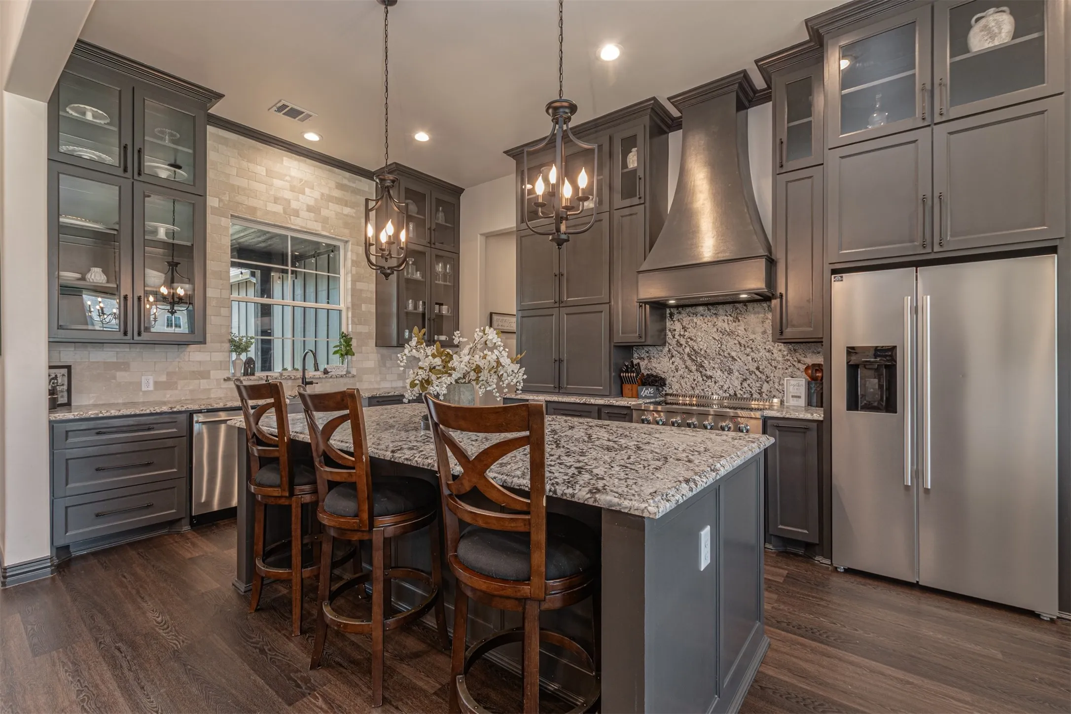 Kitchen featuring an expansive island with granite countertops, rich wood-finish flooring, dark gray cabinetry, stainless steel appliances, and a tile backsplash