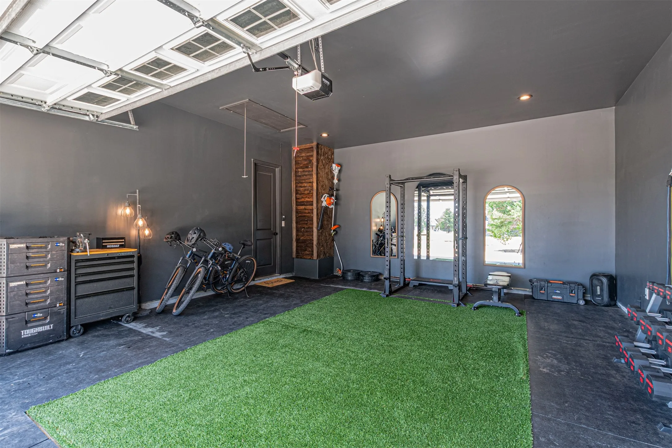 Spacious multi-purpose room featuring a paneled garage door, high ceilings with recessed lighting, and a durable flooring system combining black rubber matting with an artificial turf section
