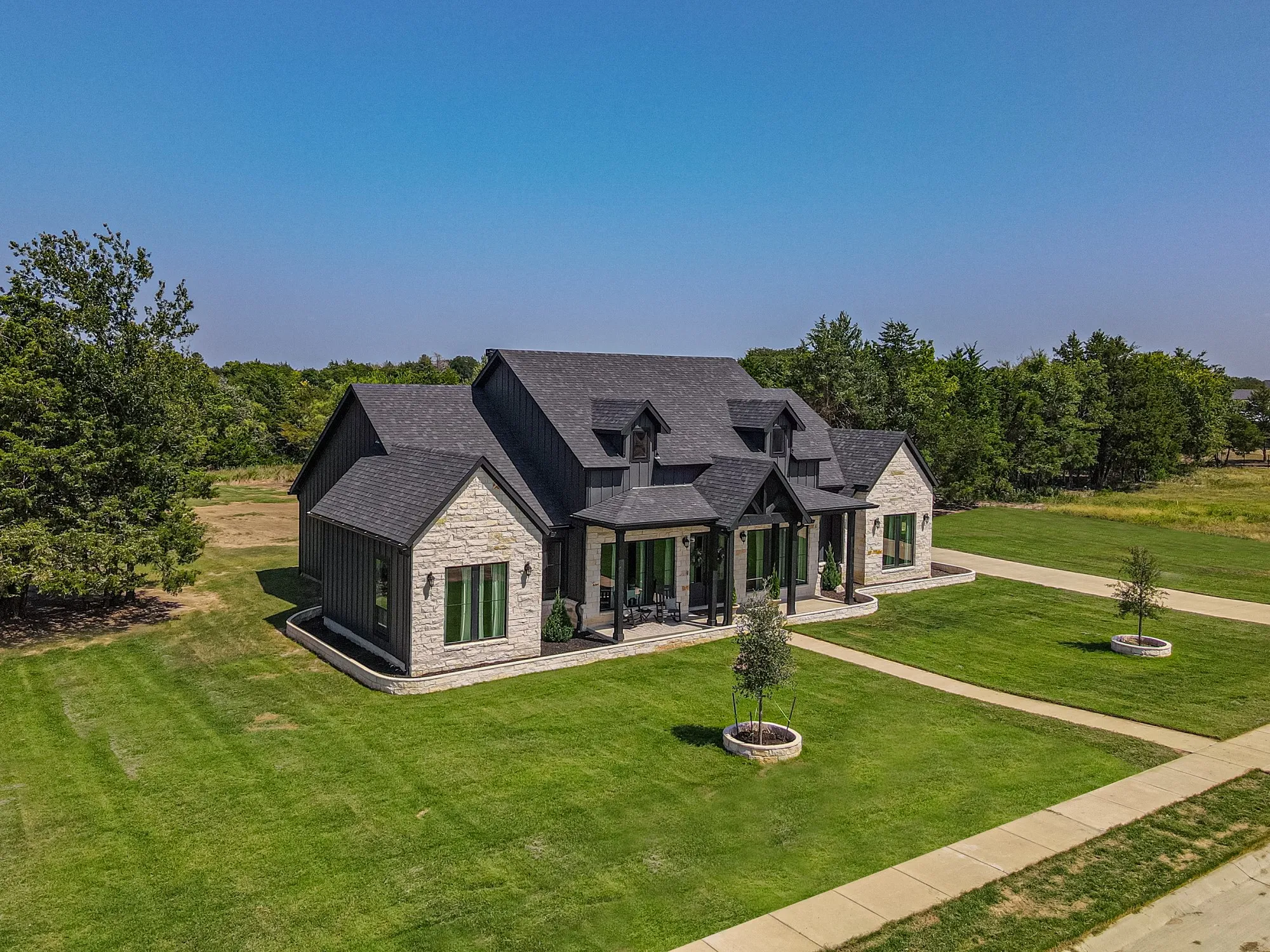 Modern farmhouse architecture featuring a stone and vertical siding exterior, a dark shingle roof, a covered front porch with dark columns, and a concrete sidewalk