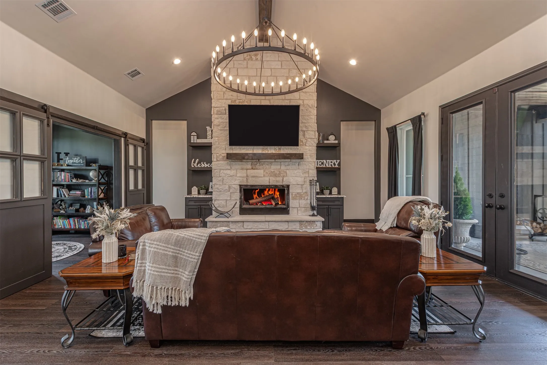 Living space featuring a stone-clad fireplace with wood mantle, vaulted ceiling with exposed wood beam, large chandelier, and dark wood-finish flooring