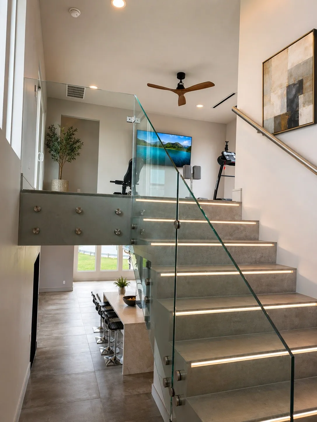 Striking modern staircase featuring glass railings and integrated LED step lighting, leading to the upper-level fitness area. Clean architectural lines and contemporary finishes create a dramatic focal point.