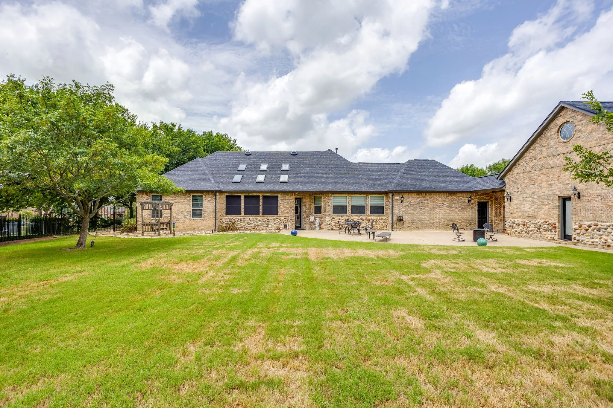 Expansive green space featuring a large patio, brick and stone exterior, multiple skylights, and mature trees