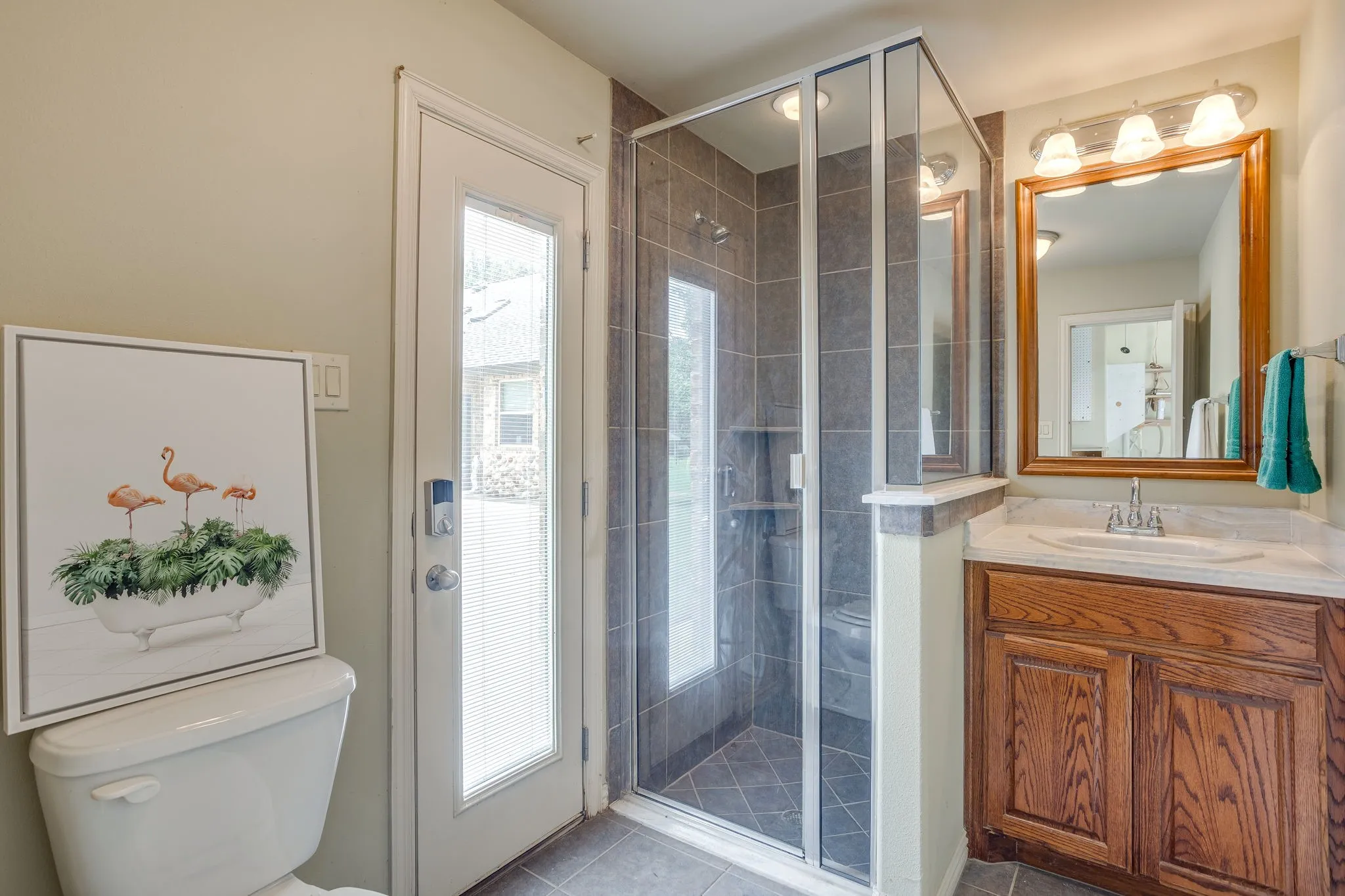 Garage Bathroom featuring a glass-enclosed shower with tile surround, a wood-finish vanity with an integrated sink and countertop, and a framed mirror with overhead vanity lighting
