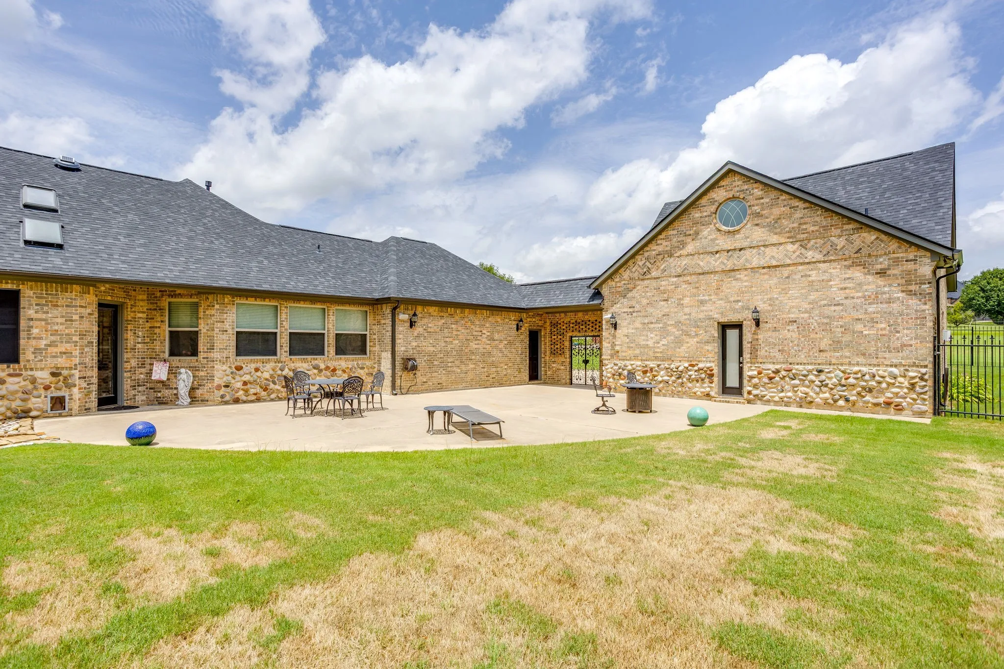 Expansive concrete patio bordered by a manicured lawn, featuring a brick and stone exterior with multiple windows and exterior doors, a black shingle roof, and a black metal fence