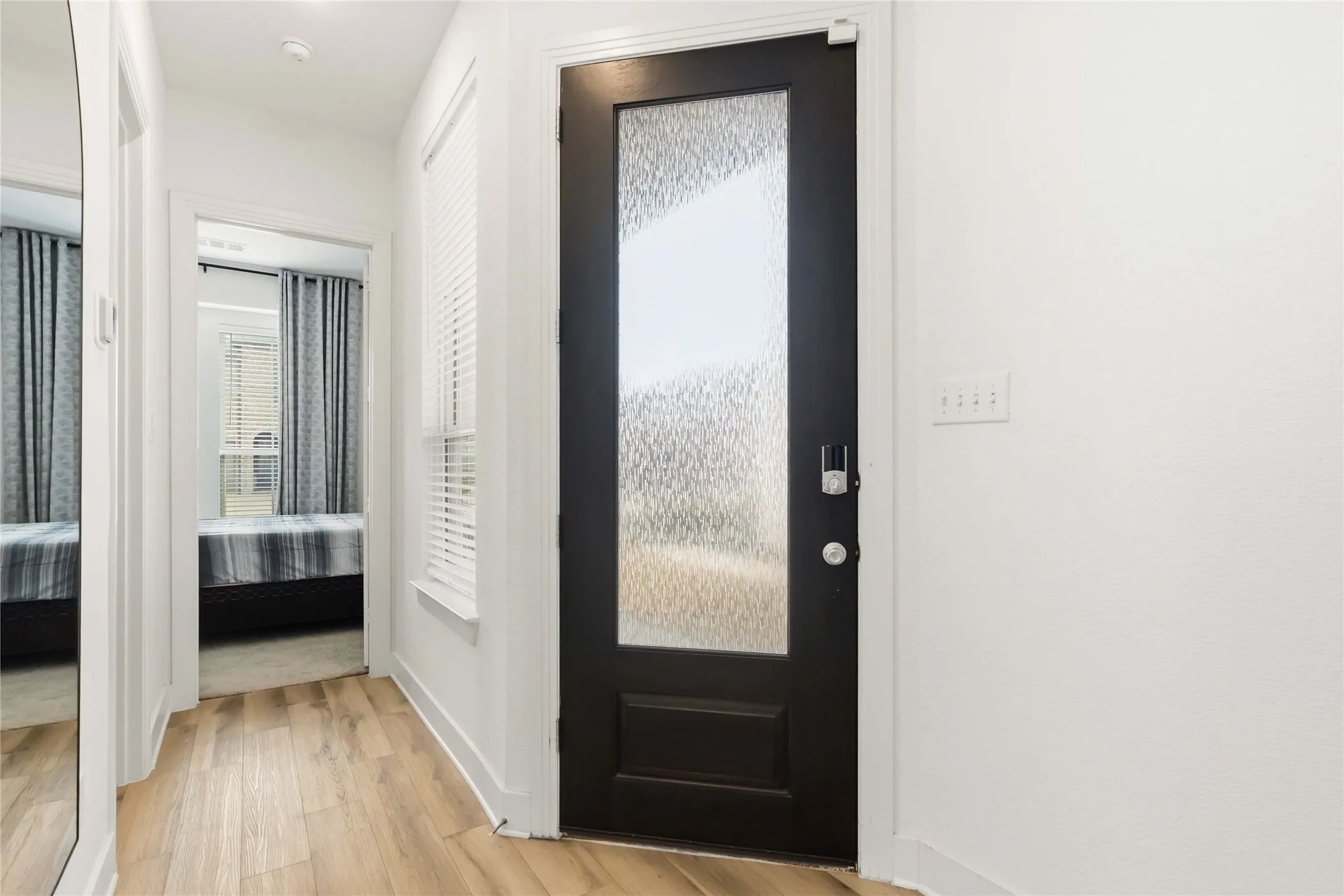 Entryway featuring a dark paneled door with a textured glass insert, wood-finish flooring, white baseboards, and a window with horizontal blinds