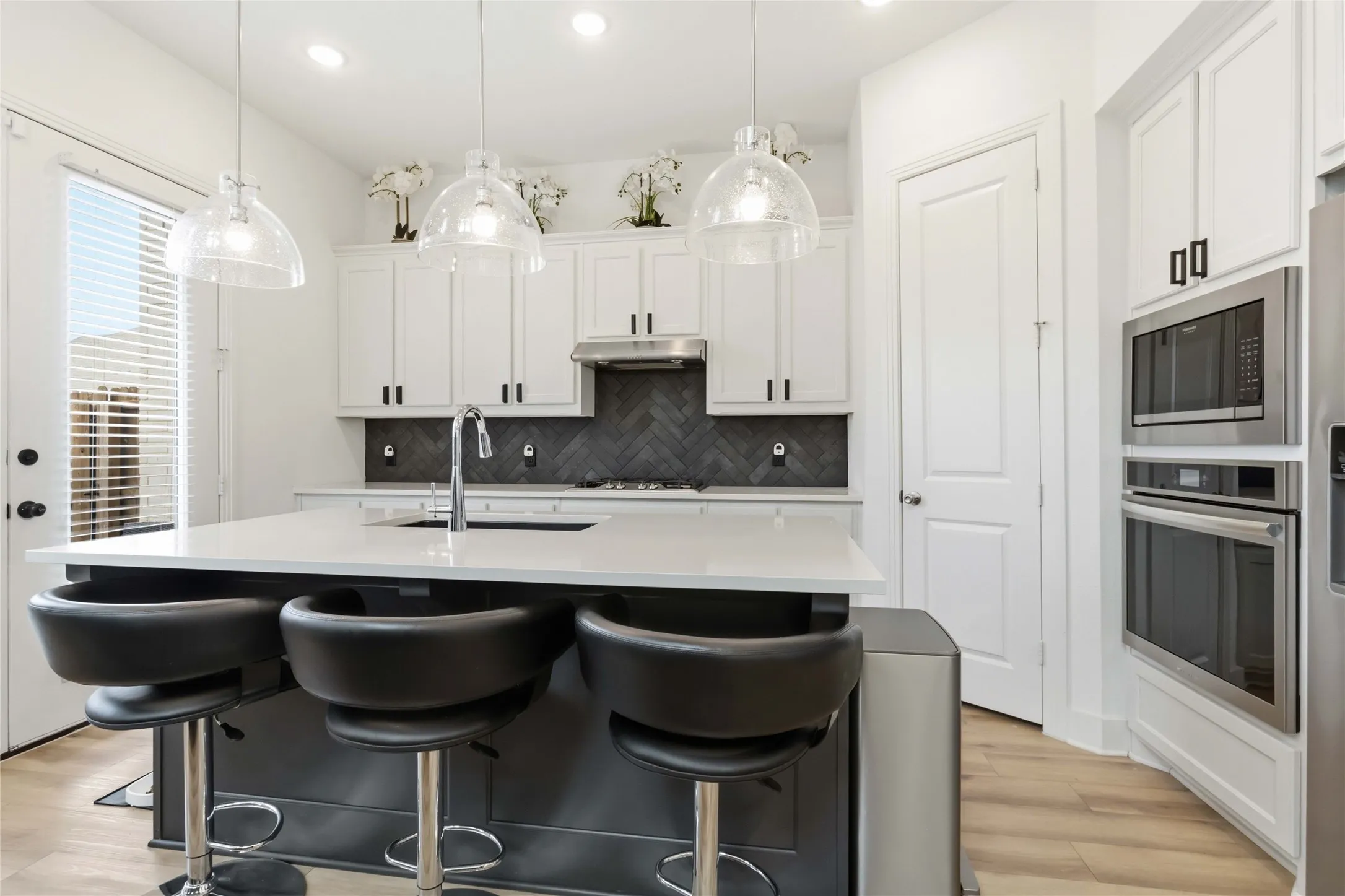Kitchen featuring a large island with a light-toned countertop, white cabinetry with dark hardware, a dark herringbone tile backsplash, stainless steel appliances, and wood-finish flooring