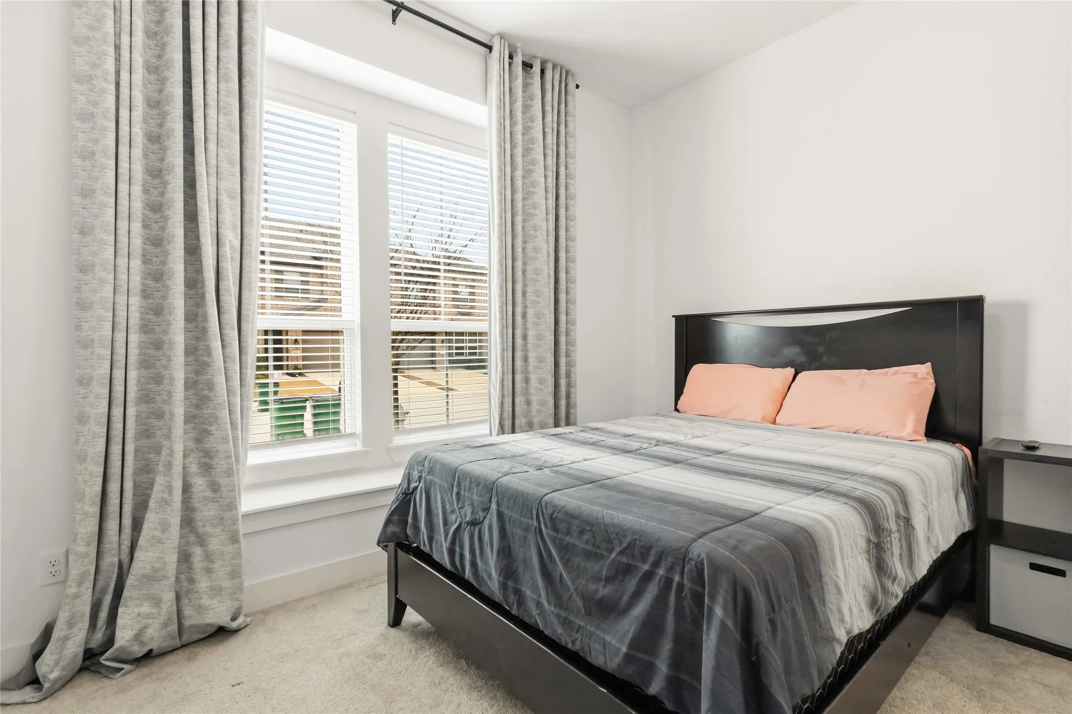 Bedroom featuring light-colored carpet flooring, white walls, and a large window with blinds and patterned drapes
