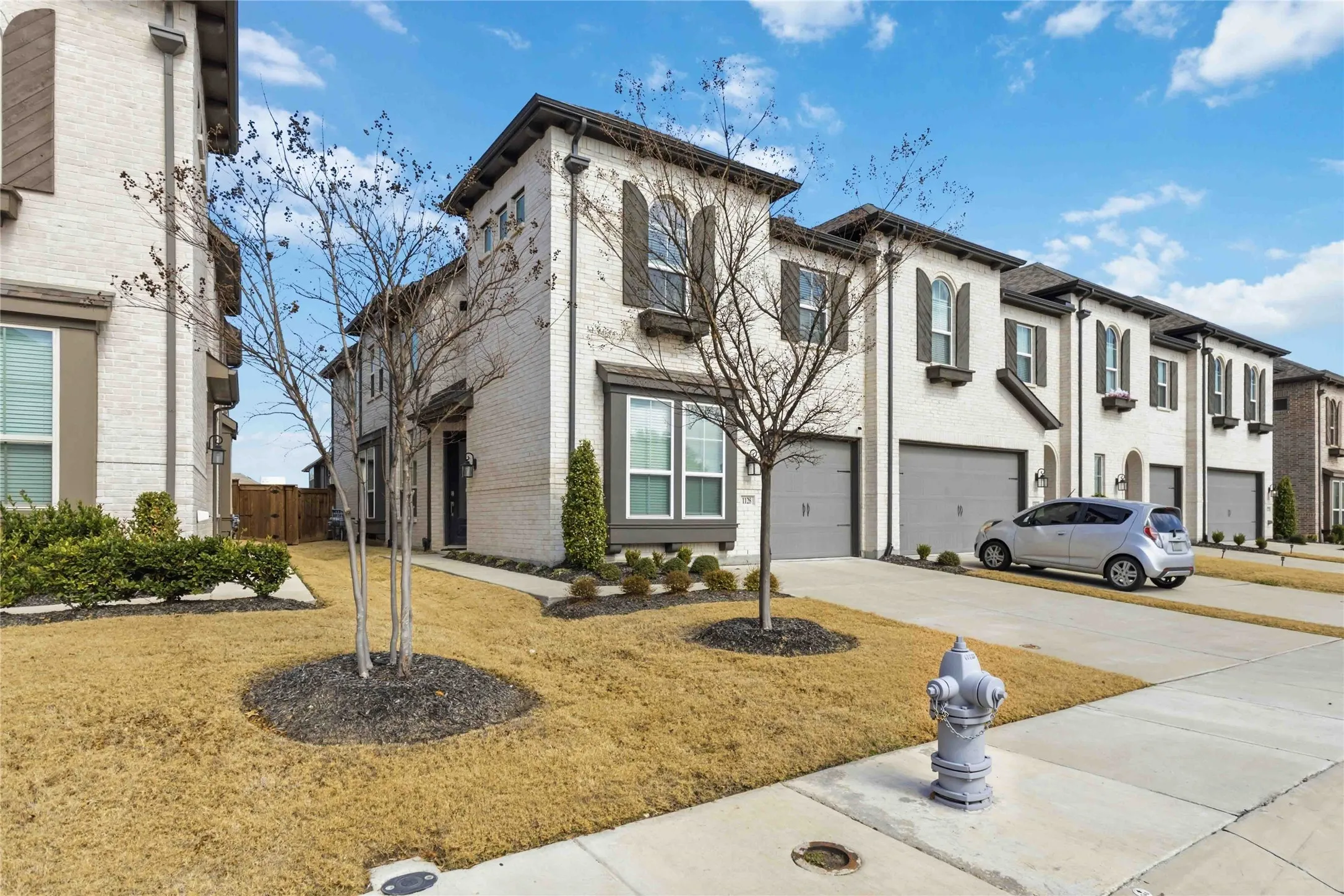 Contemporary townhome exterior featuring light-colored brick, dark window shutters, arched window accents, a dark entry door, and an attached two-car garage